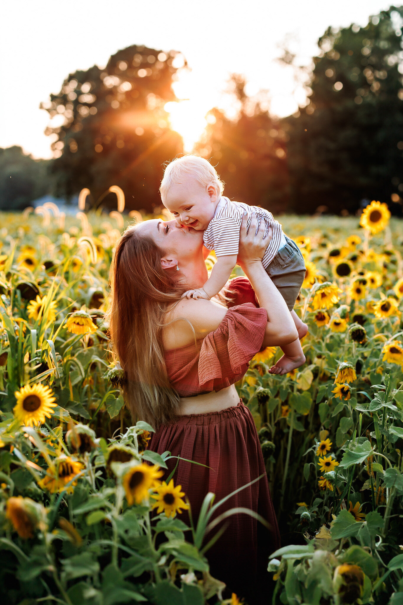 A mother surrounded by a field of sunflowers, holds her toddler son up and gives him a sweet kiss on the cheek