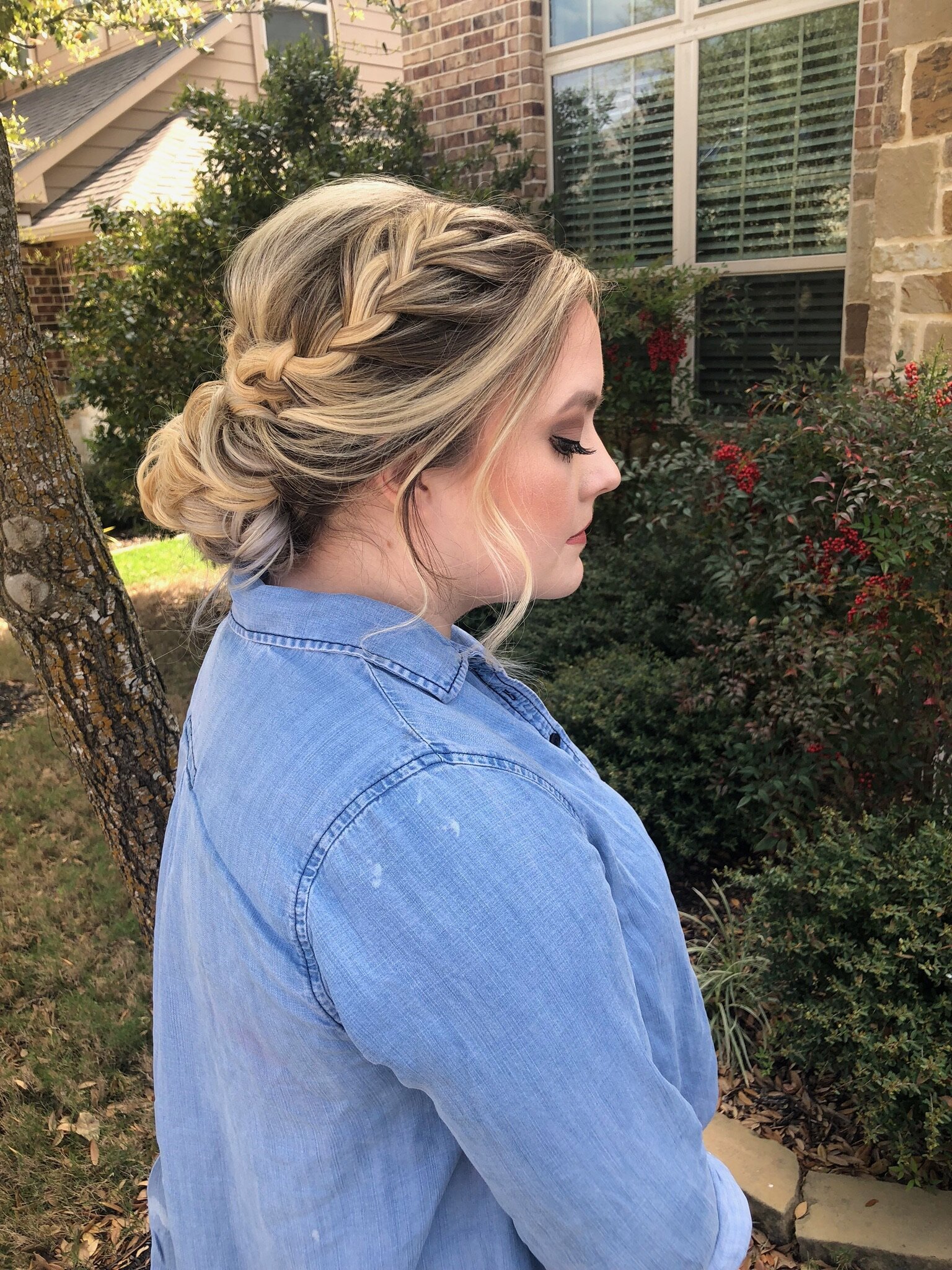 Woman with a braided updo in a denim shirt stands outdoors near greenery, reflecting on the enchanting bridal beauty experience Texas offers.
