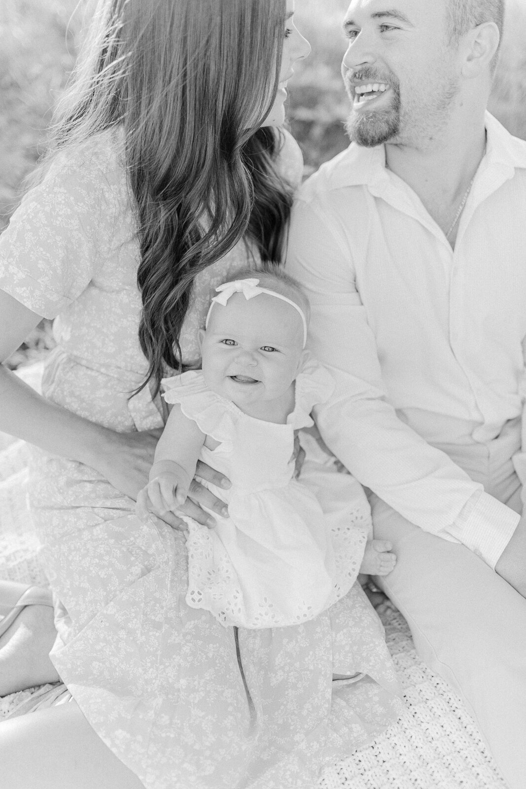 A black and white photo of a mom and dad holding their baby girl in their lap during photo session with Boston family photographer Corinne Isabelle