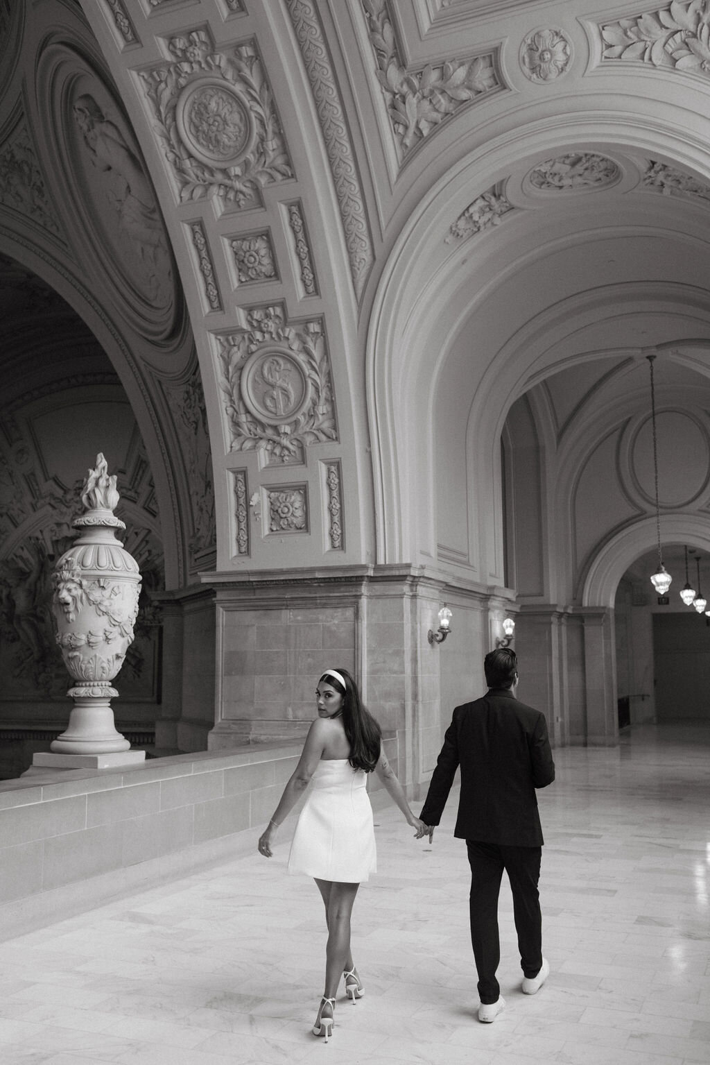 A couple holding hands walks through an ornate hallway in San Francisco City Hall, with the woman in a white dress looking back and the man in a suit leading forward.