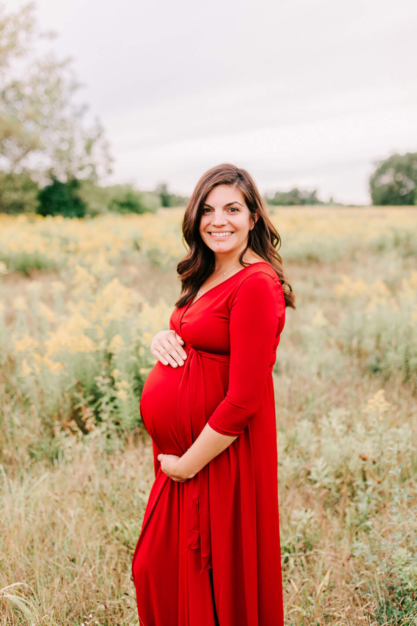 expecting mother of 3 stands in a red dress and holds her belly in a beautiful field of green and yellow.