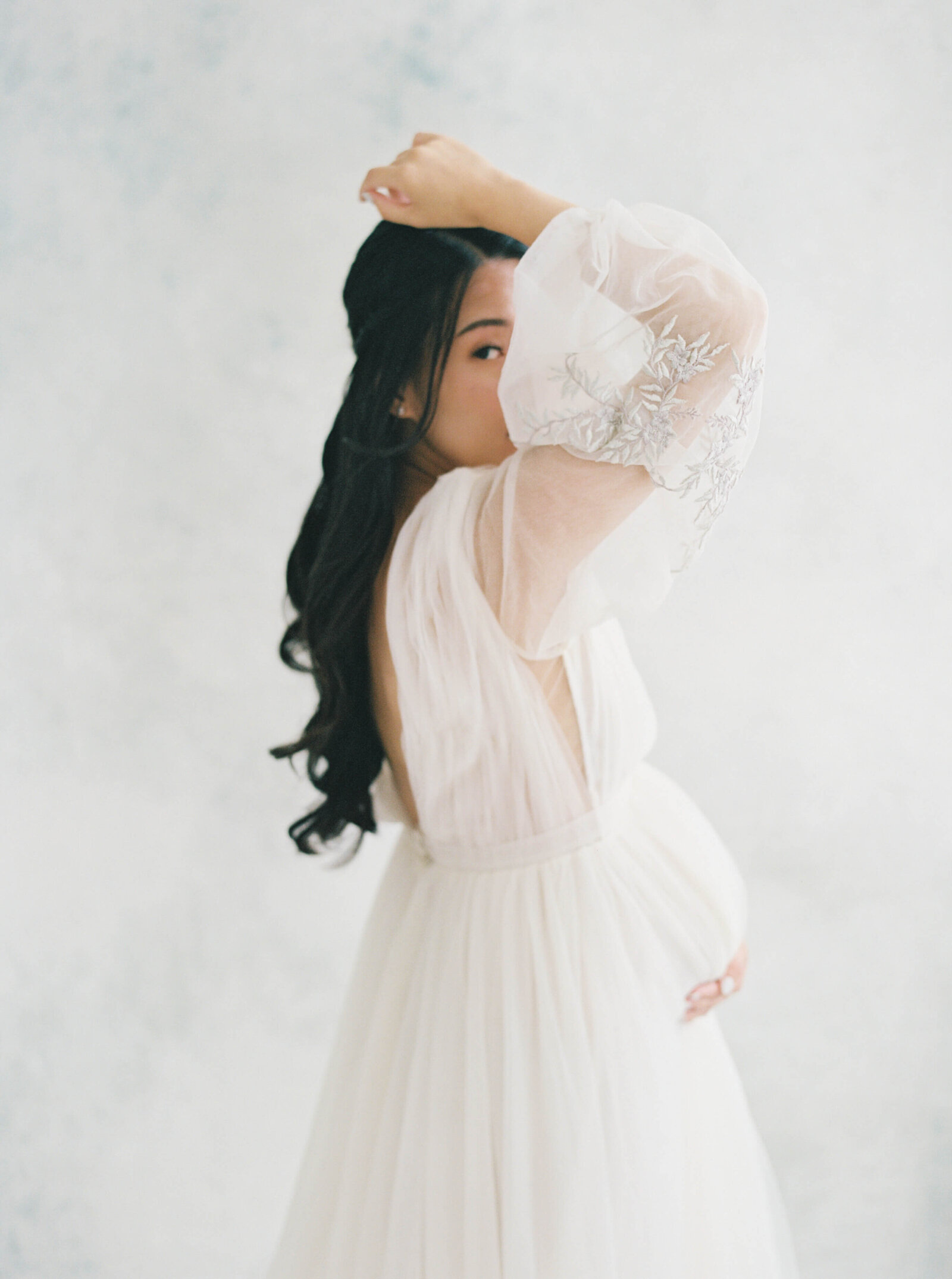 A mother-to-be in a classic white dress, gently cradling her belly in a softly lit studio.