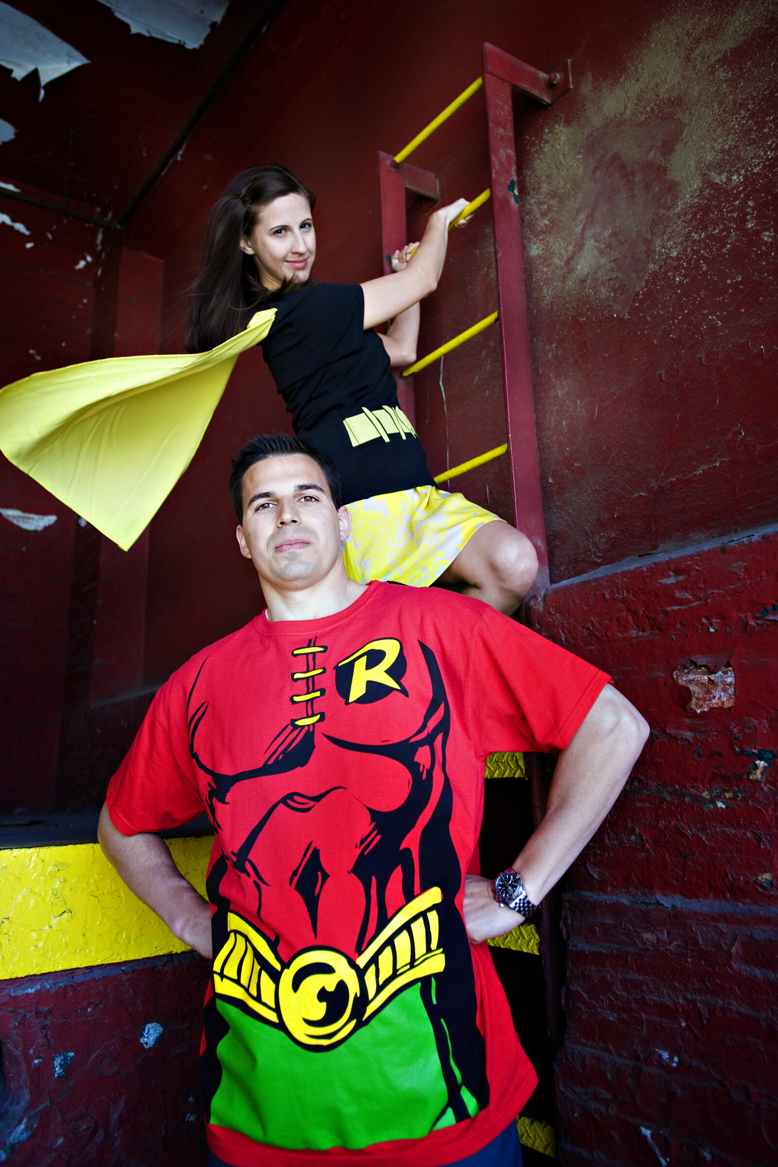 A couple in superhero outfits standing together as one climbs up a ladder 