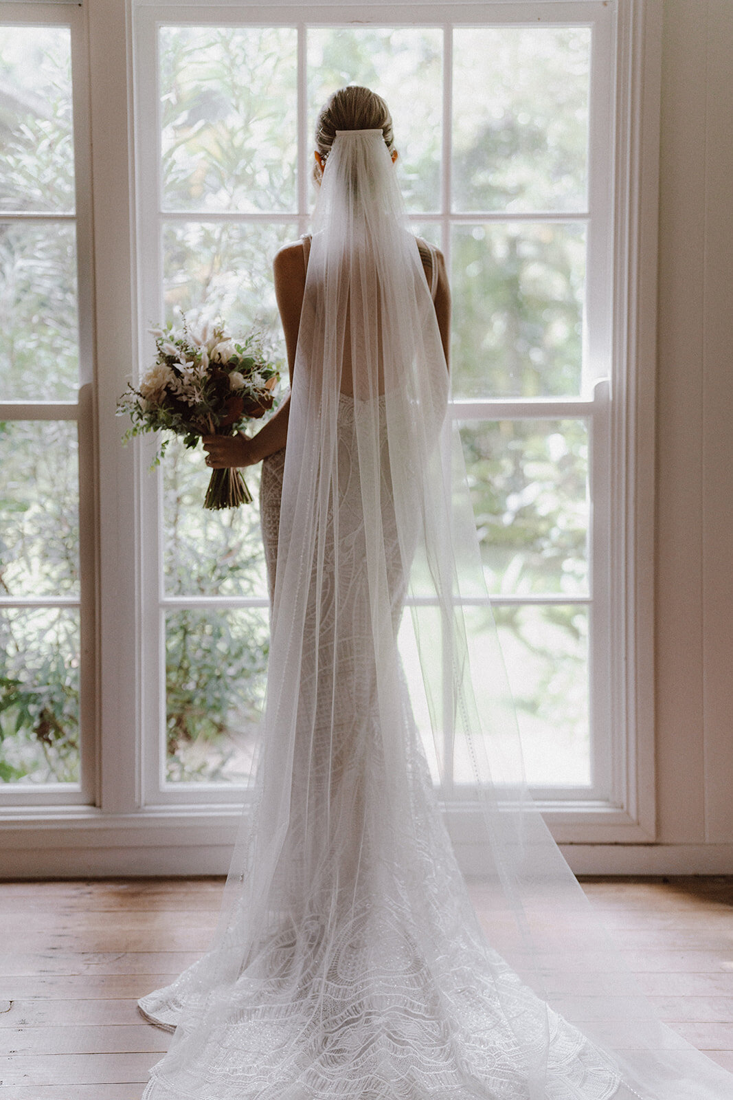 Bella Rae Bride Heather looking out the window at Tiffanys Maleny With a long Veil attactched to a low twisted bun captured by Artography Weddings