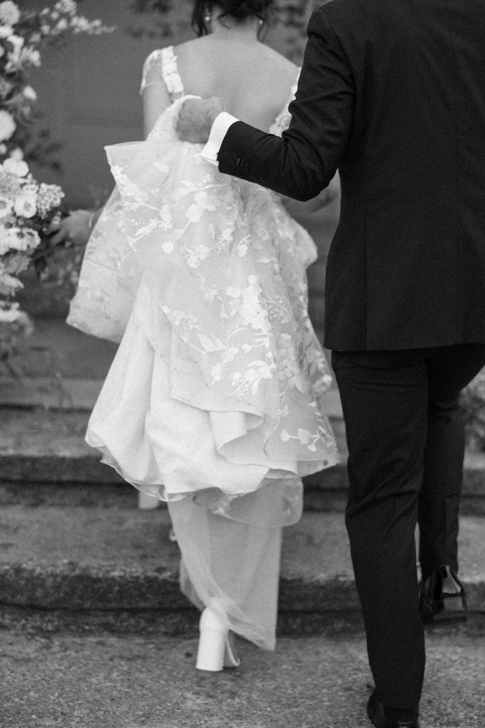 Candid black & white photo of groom holding bride's floral lace train as they walk up stone garden steps 