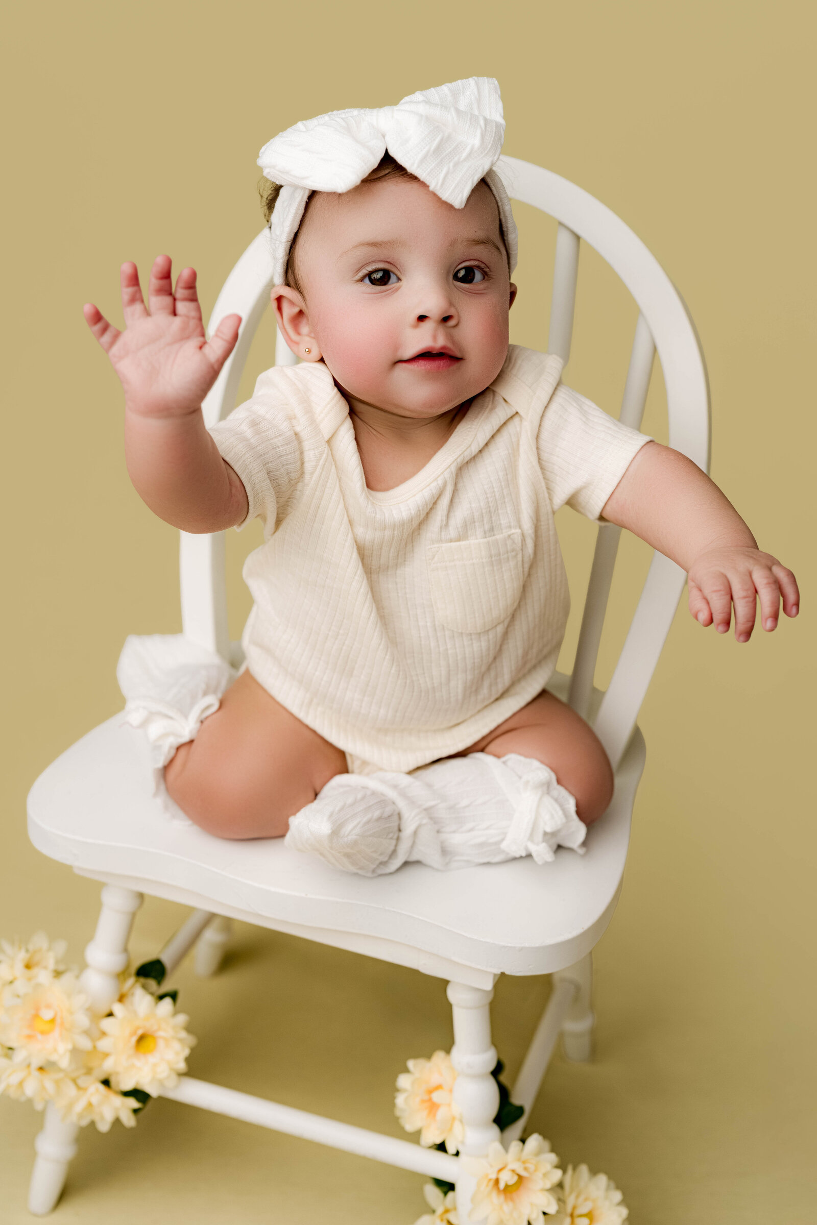 Baby girl sitting on a white chair with fresh daisies, reaching up during a whimsical milestone session in Waxhaw, NC.