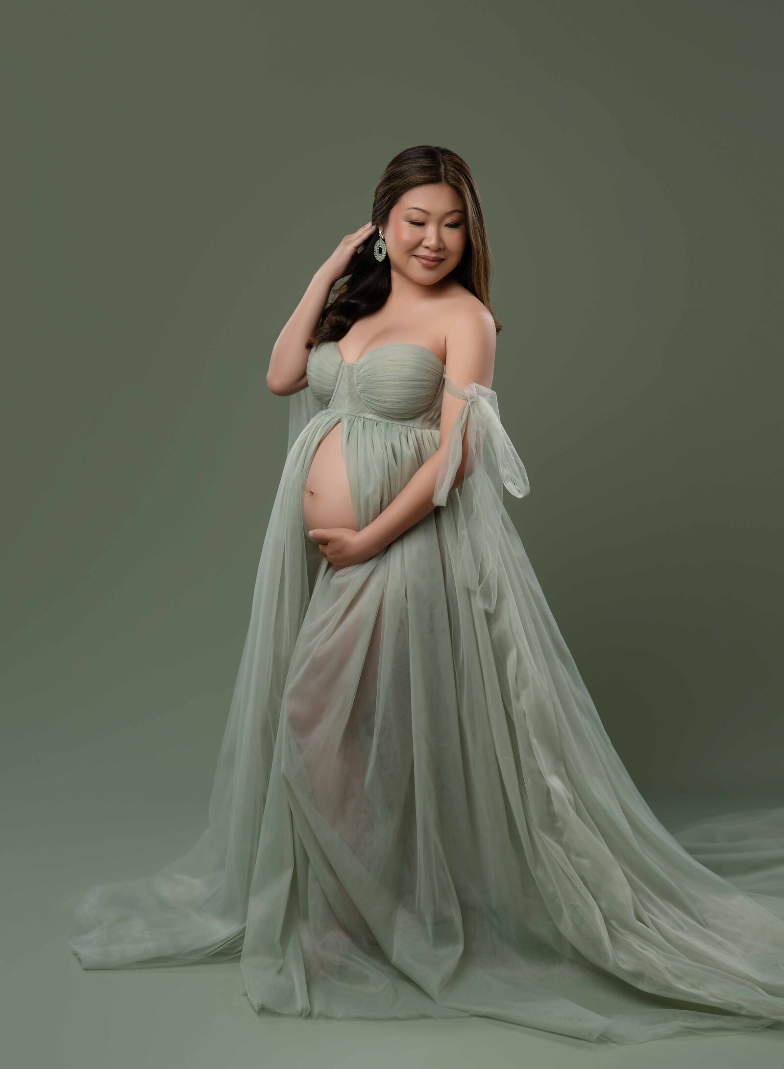 Pregnant women wearing sage mint tulle maternity dress, holding her baby bump and looking over shoulder.
