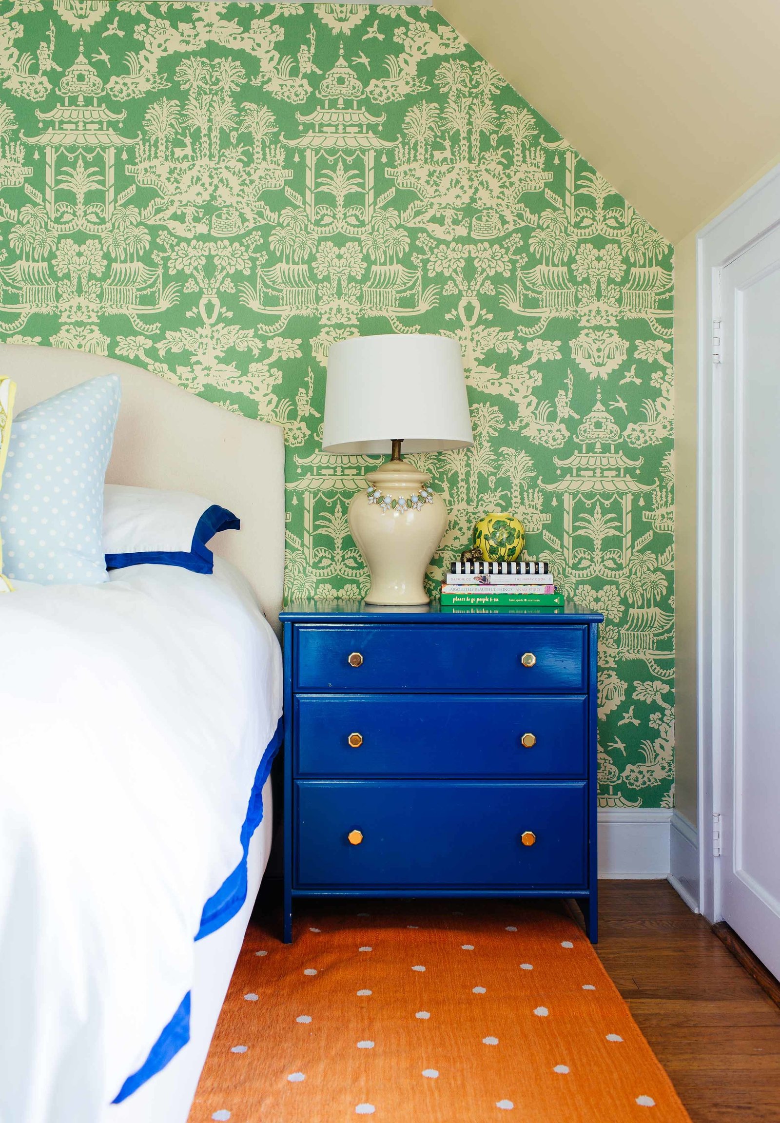 A white bed and blue end table in front of a green wall papered wall.
