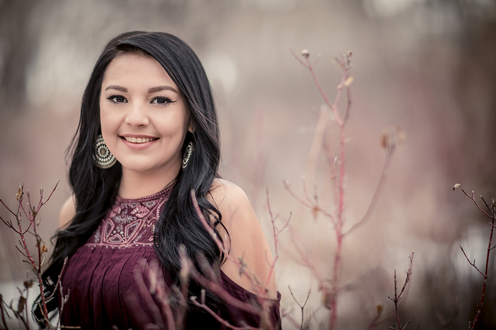 High School pictures. Girl smiling, black hair with maroon shirt with complimentary color with red twig dogwood trees at River Front Park in Billings, MT.