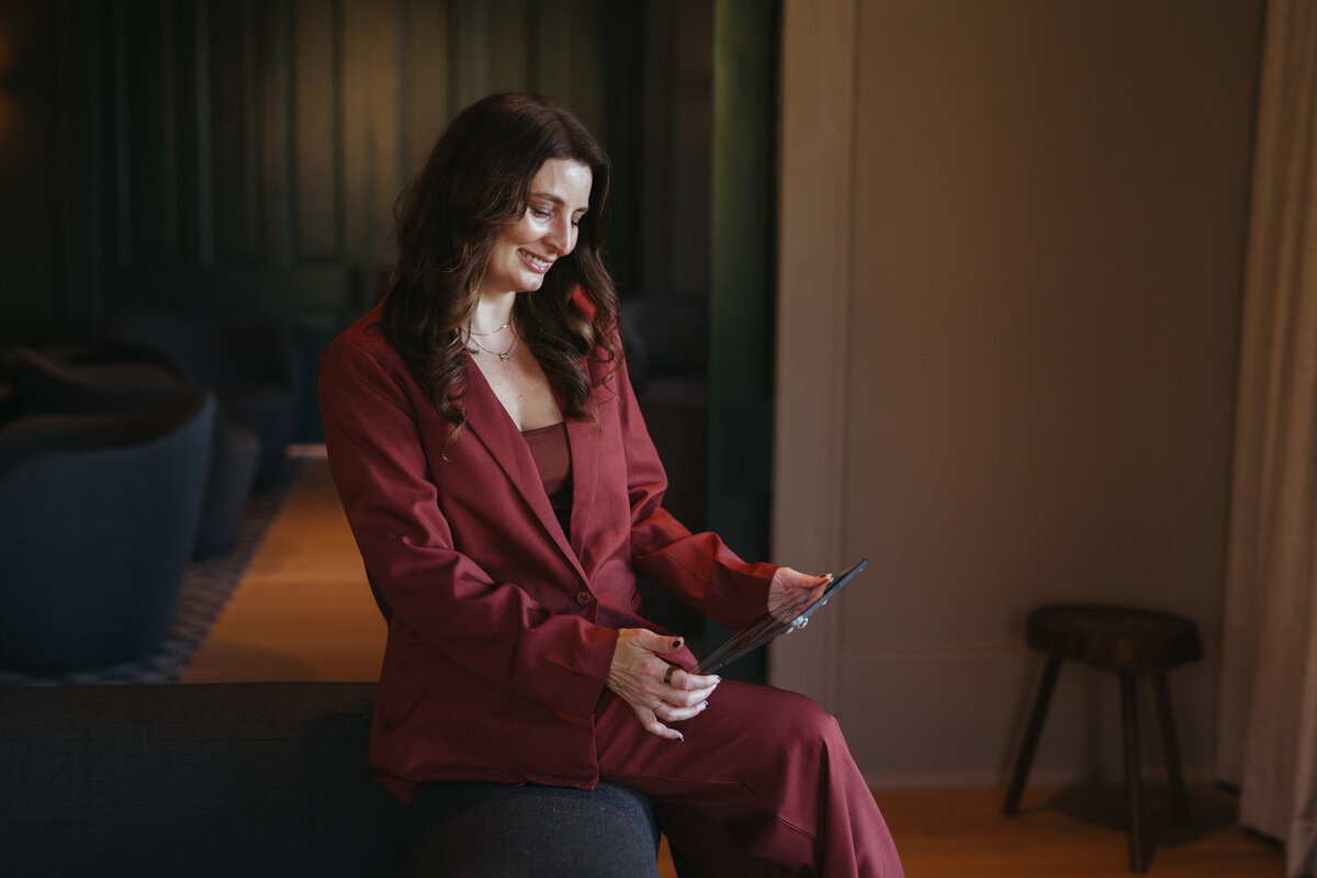 A woman in a maroon suit sits on a sofa, smiling at her tablet; dark wood panels form the background, evoking an executive retreat planning ambiance.