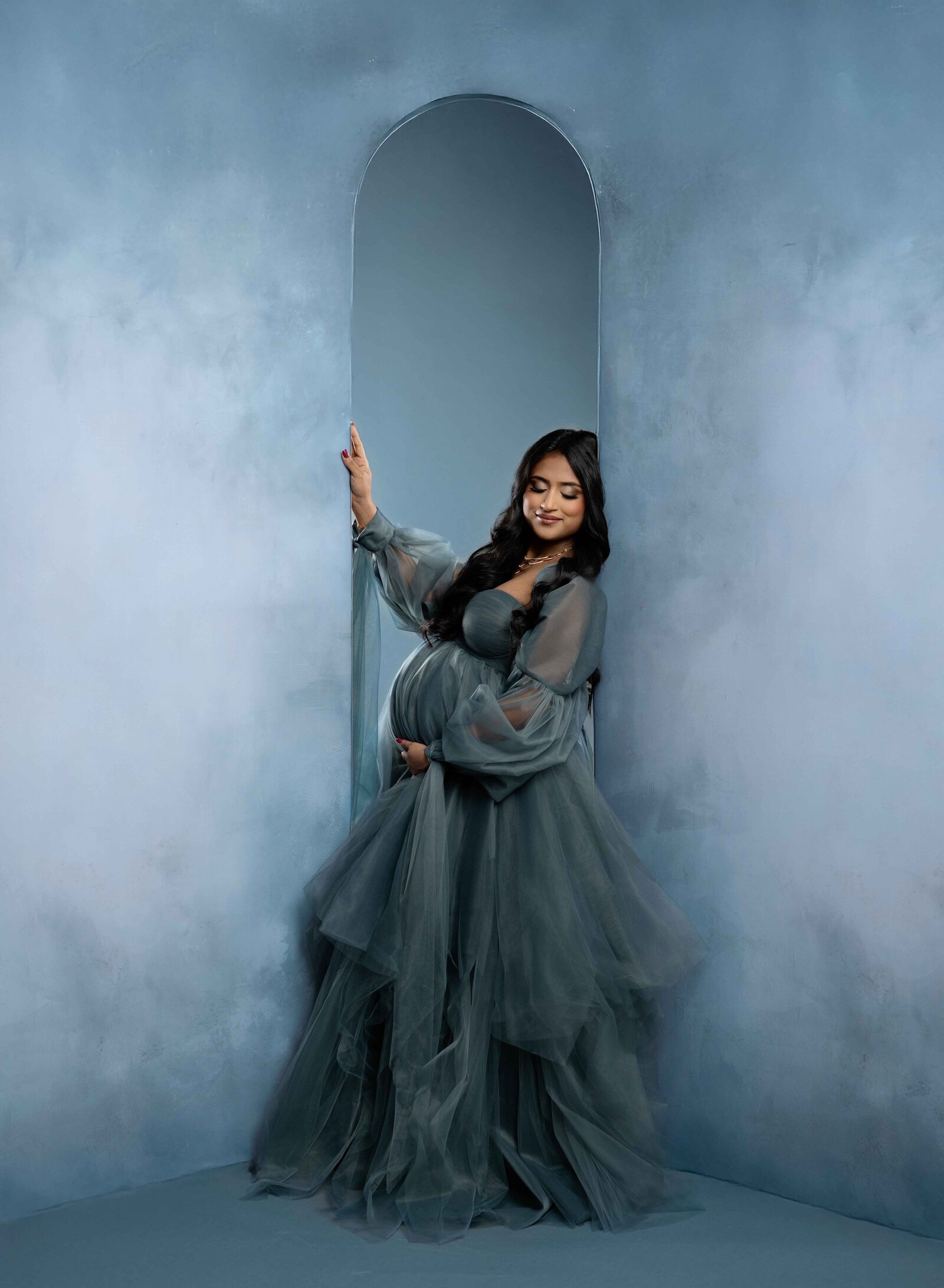 Pregnant women leaning on blue arch wall and holding her baby bump.