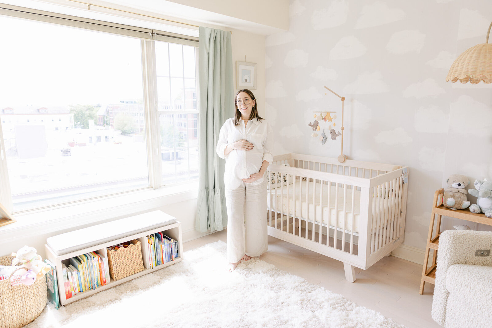 A glowing expectant mother stands barefoot in a sun-filled, cloud-themed nursery, gently holding her baby bump next to a wooden crib and a mobile of animals. Captured in soft neutrals and cozy textures, this peaceful maternity photo documents the beauty of new beginnings for families who value slow moments, dreamy light, and heartfelt connection.
