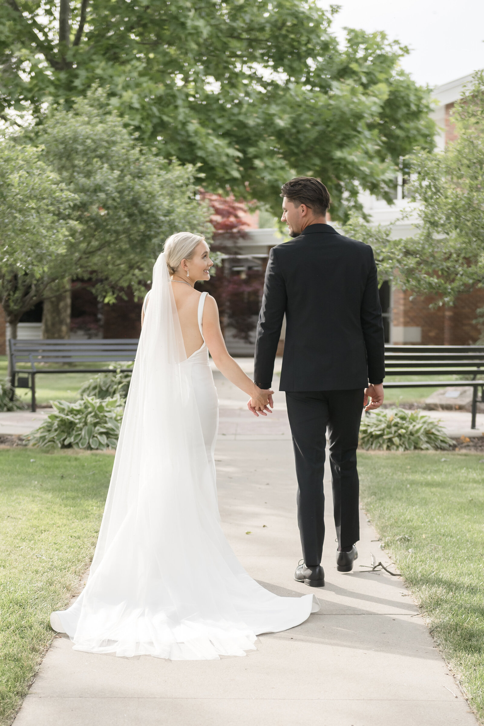 couple holding hands walking on Alma College Campus while bride looks over her shoulder