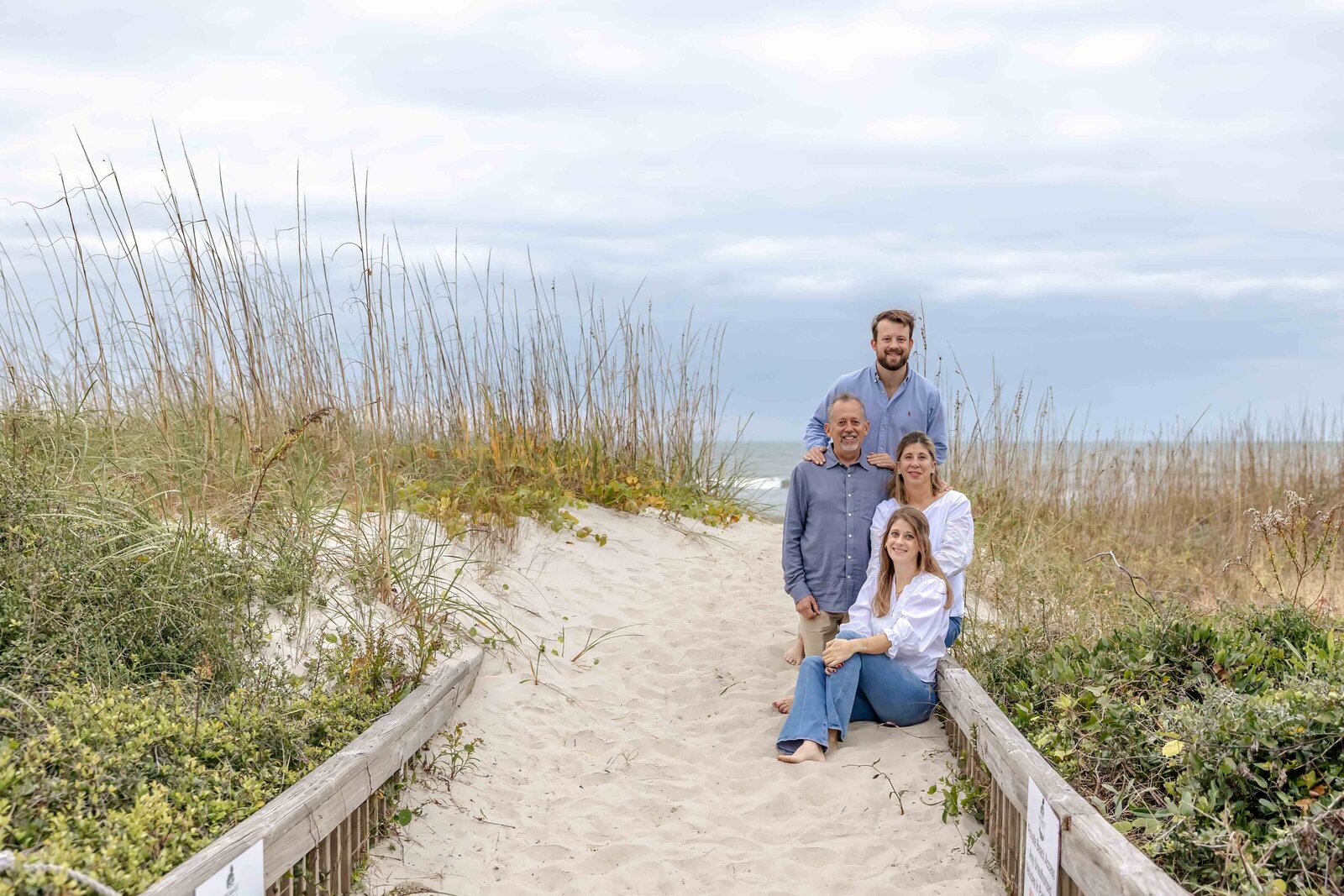 A family sits on a sandy path lined with sea oats, leading to the beach. Dressed in white and blue, they enjoy a tranquil coastal setting, ideal for destination family photographer NC, capturing serene and natural family moments.