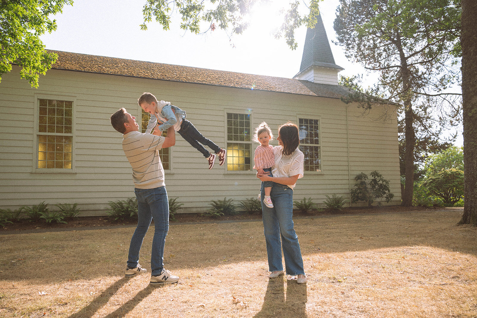 Parents play with their two children in the sunshine, lifting and holding them outside a white building during family photos in Irvine, CA.