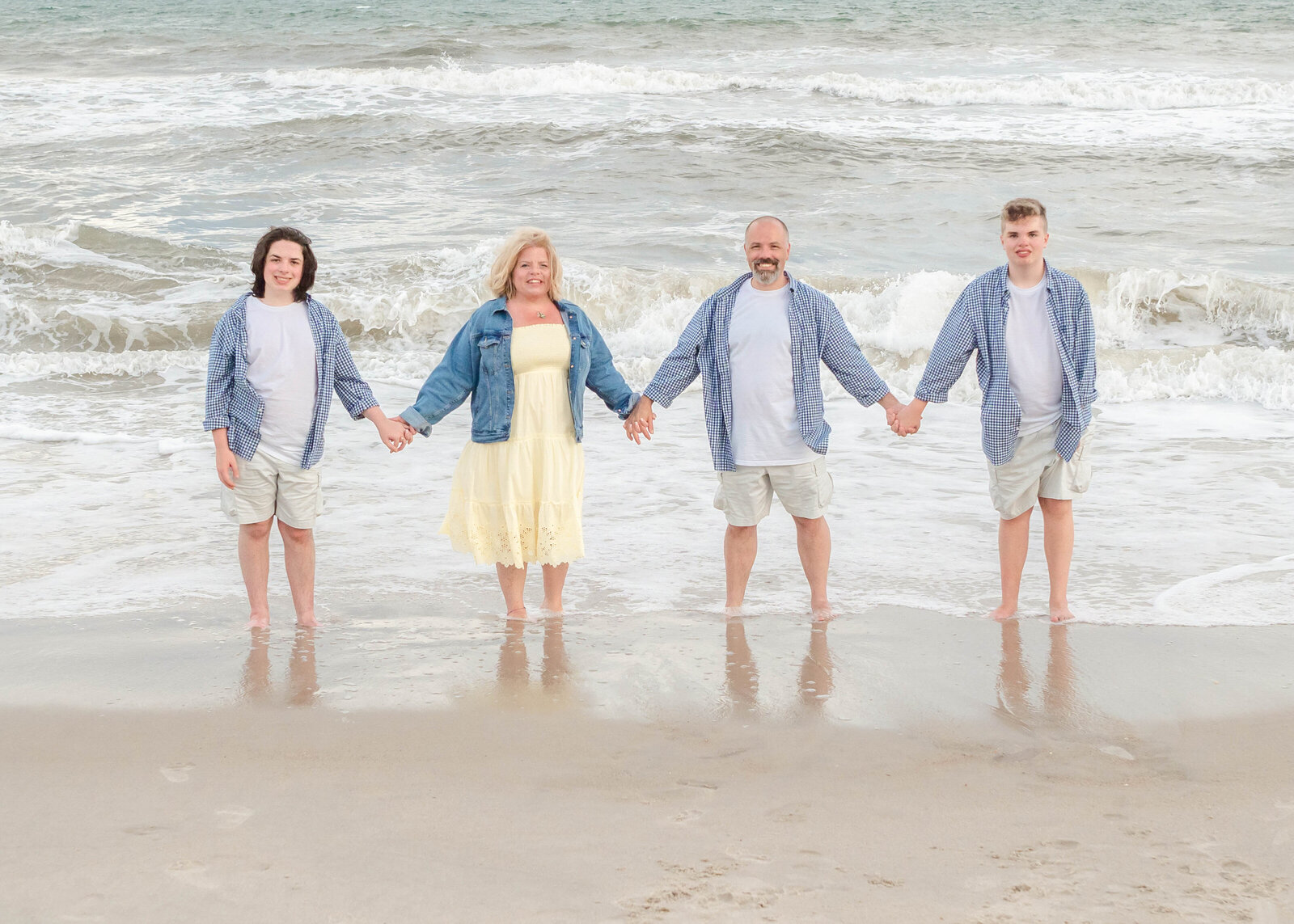 A family of four stands in the surf at North Topsail Beach, holding hands and wearing matching blue plaid shirts and white shorts. The mother stands out in a yellow dress, adding a cheerful touch. This image captures the essence of North Carolina family photography, ideal for beachside memories.