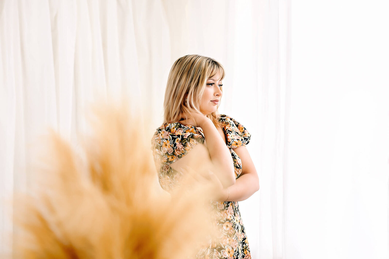 A woman with blonde hair stands in a brightly lit room, wearing a floral dress. She gazes thoughtfully to the side, with her hand gently touching her neck. In the foreground, there is a blurred cluster of wheat or dried flowers.