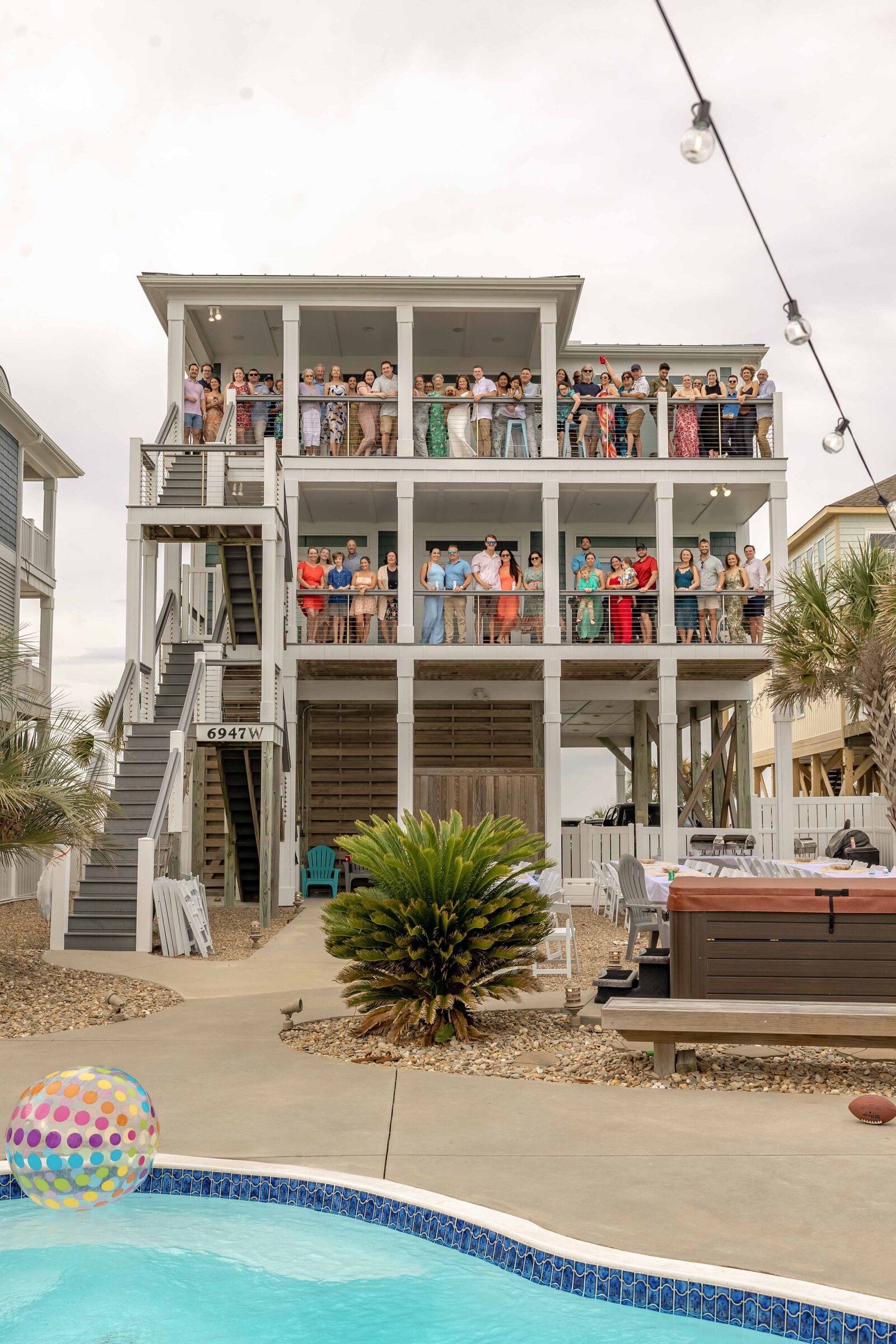 A vibrant group of wedding guests gathers on the balconies of a beach house in Topsail Beach, NC, celebrating love and coastal charm, with a pool and playful decor adding to the fun atmosphere.