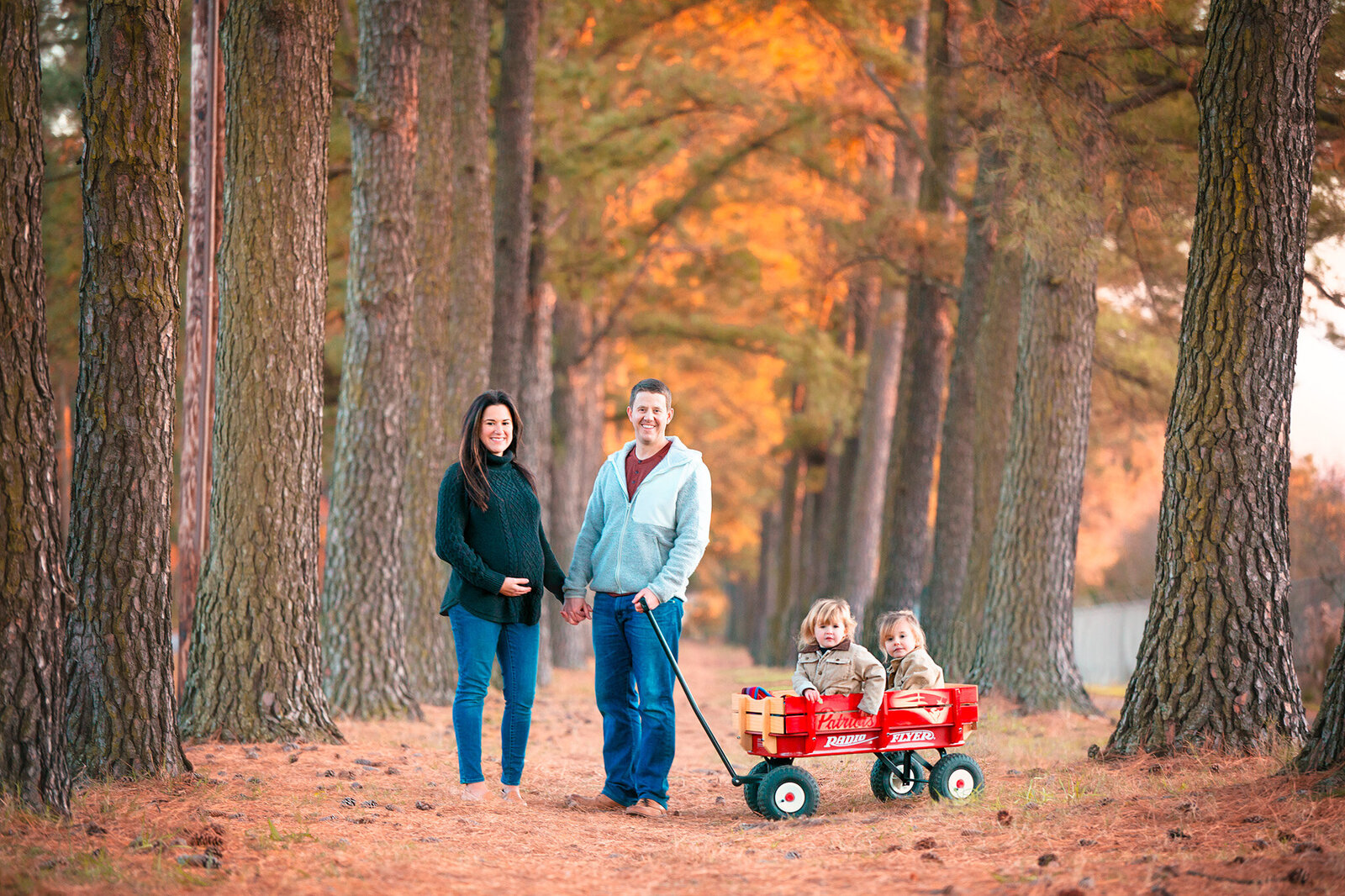 Family photographer Hampton Roads va photographs outdoor family photos with parents holding hands as the mother holds her pregnant stomach and their children sit in a red wagon