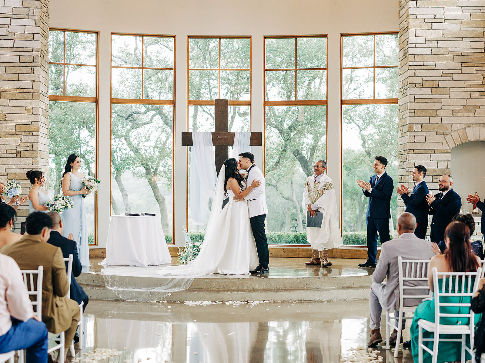A bride and groom share their first kiss at the altar during a wedding ceremony in a bright, windowed chapel at Canyonwood Ridge in Austin, surrounded by their wedding party and guests applauding.