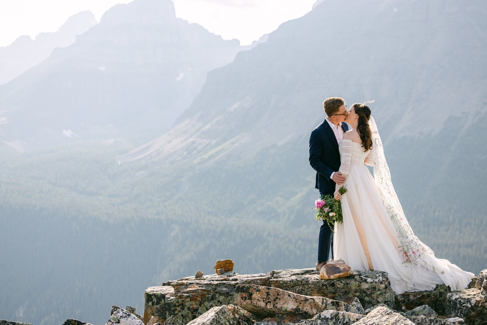 A breathtaking moment as a couple stands atop a mountain in Banff National Park, the bride’s dress flowing in the wind on a warm summer day. Arriving by private helicopter for their intimate ceremony, this dramatic photograph by the Canadian Rockies best elopement photographer captures the essence of adventure, love, and epic romance in the Canadian Rockies.