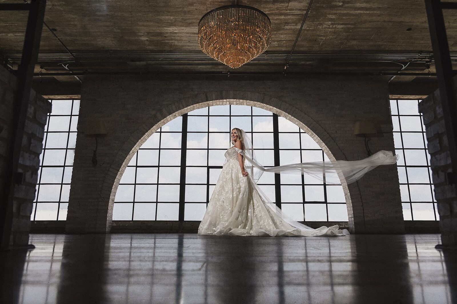 Luxury bridal portrait at The Astorian in Houston, TX by Logrono & Co. Weddings, featuring a bride in a lace gown with a dramatic flowing veil, framed by arched industrial windows and a grand chandelier.