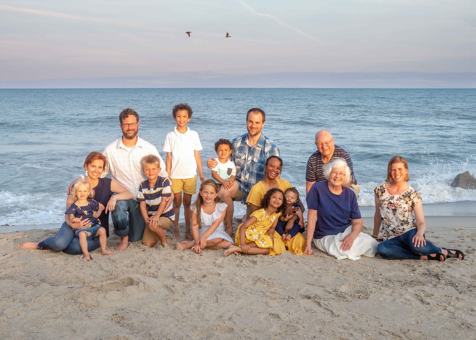 A large family gathers on the beach at sunset, with children and adults dressed in vibrant colors. The warm evening light and ocean waves create a picturesque scene, ideal for destination family photographer NC, capturing joyful family reunions.