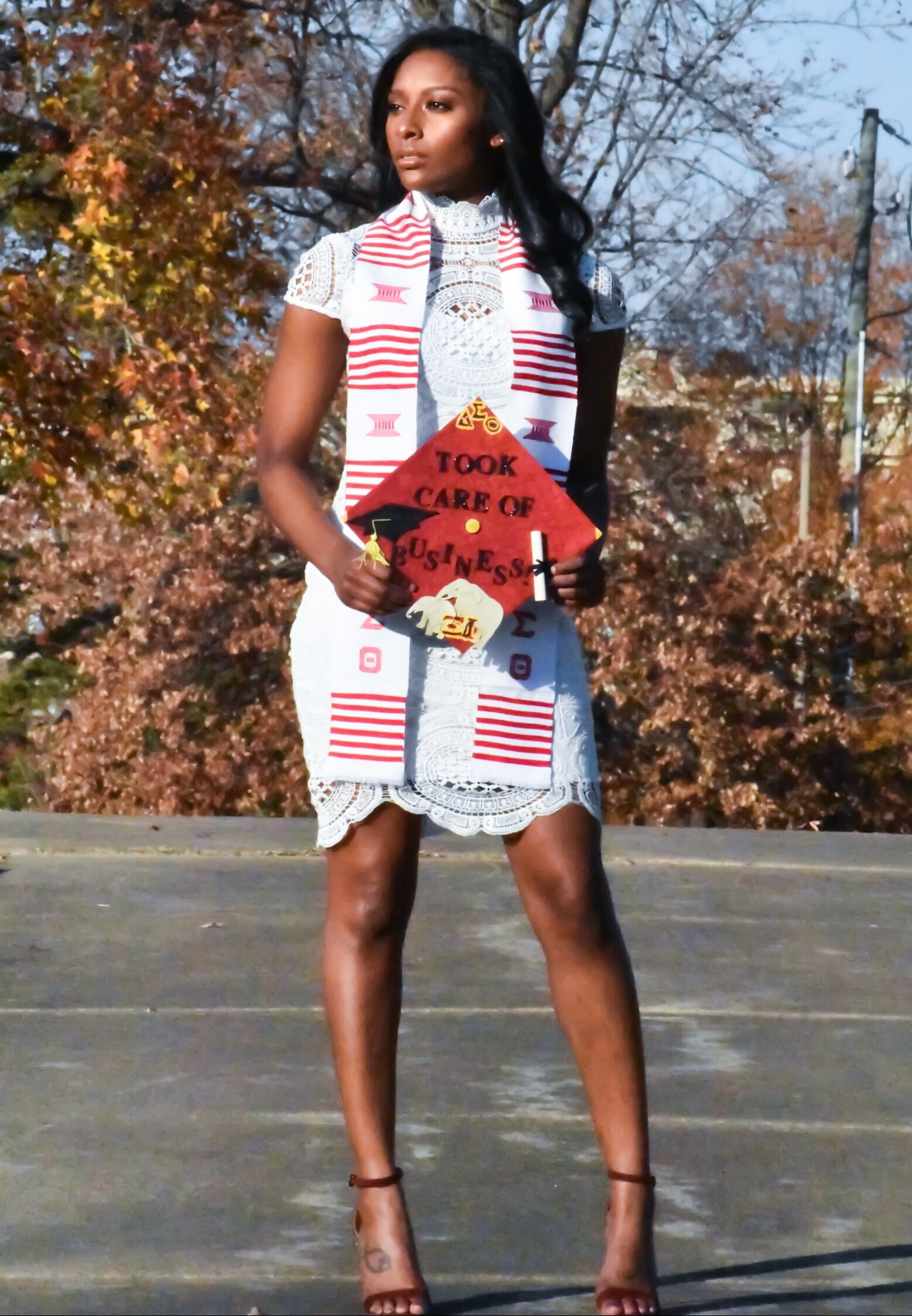 a senior dressed in a white dress with her red and white stole draped over her shoulder holding a red graduation cap. photographed by Millz Photography in Greenville, SC