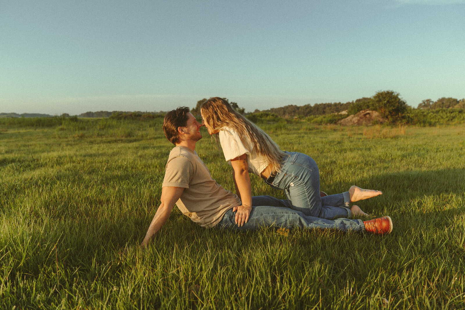 Romantic couple playfully touching noses during golden hour in a sunlit open field in Central Florida. The woman is barefoot, leaning over her partner with a candid smile, capturing natural, cinematic love and connection.