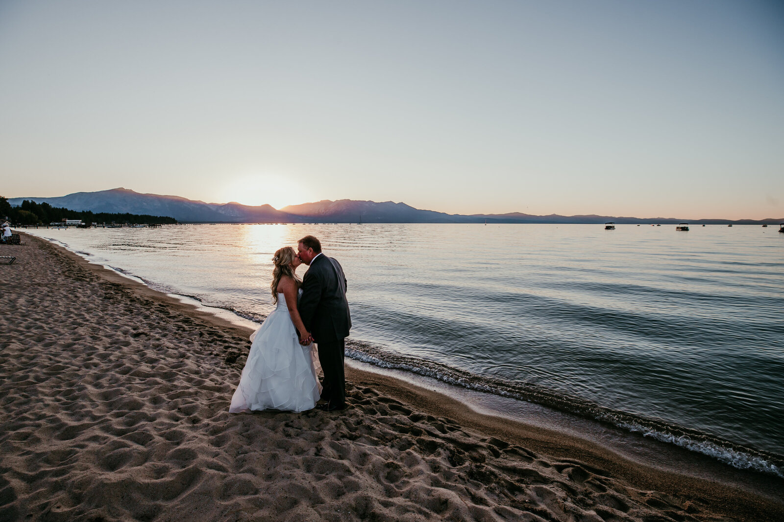 A newlywed couple shares a romantic kiss on the sandy shores of Lake Tahoe at sunset, with golden light reflecting off the calm water.