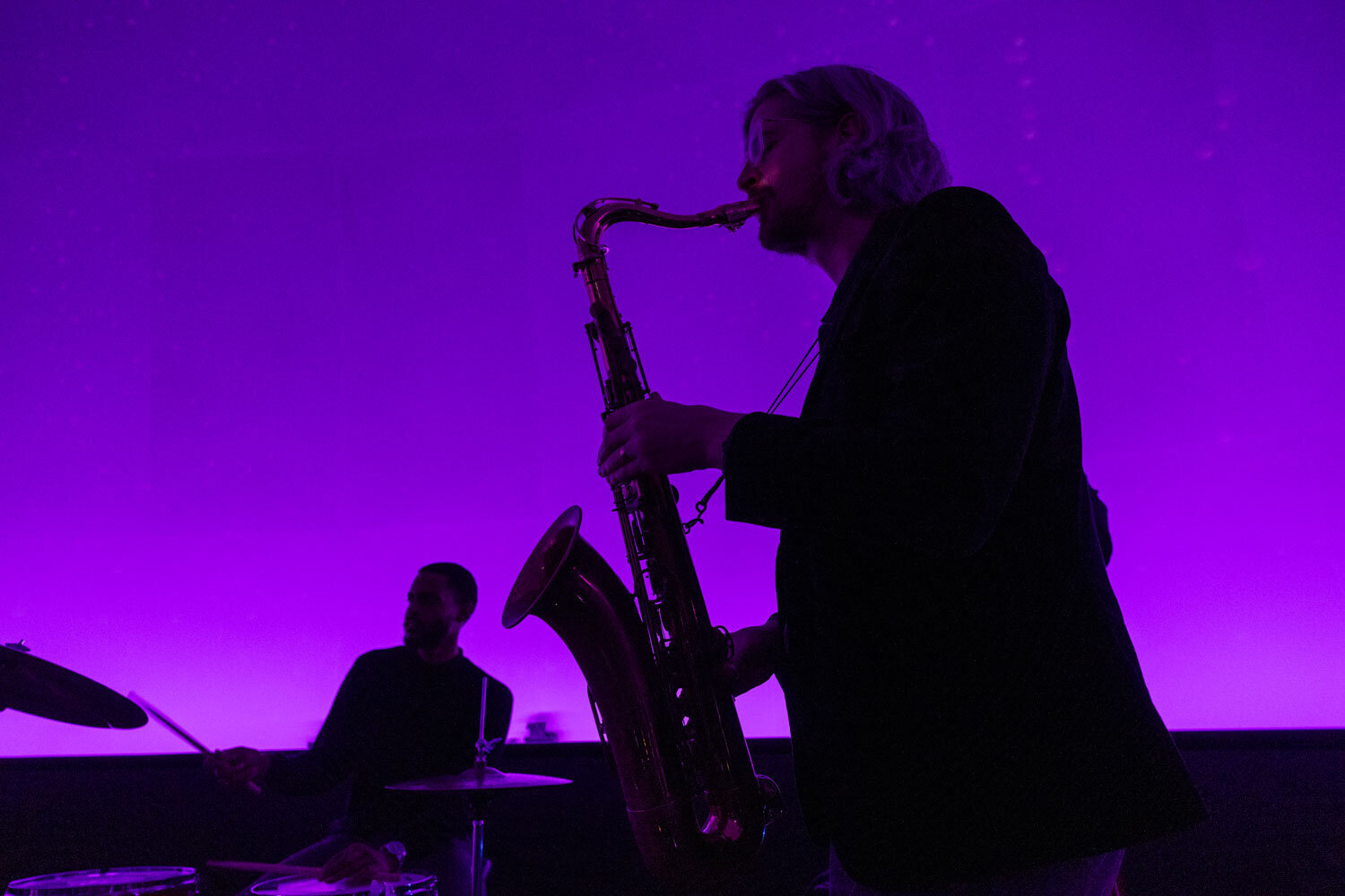 Saxophone player and drummer performing under purple lighting