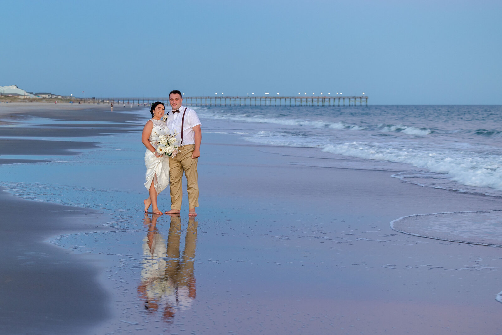 The bride and groom stand barefoot on the shoreline at sunset with the Kure Beach pier in the background, captured during a romantic Kure Beach, NC wedding, blending coastal charm and natural beauty.