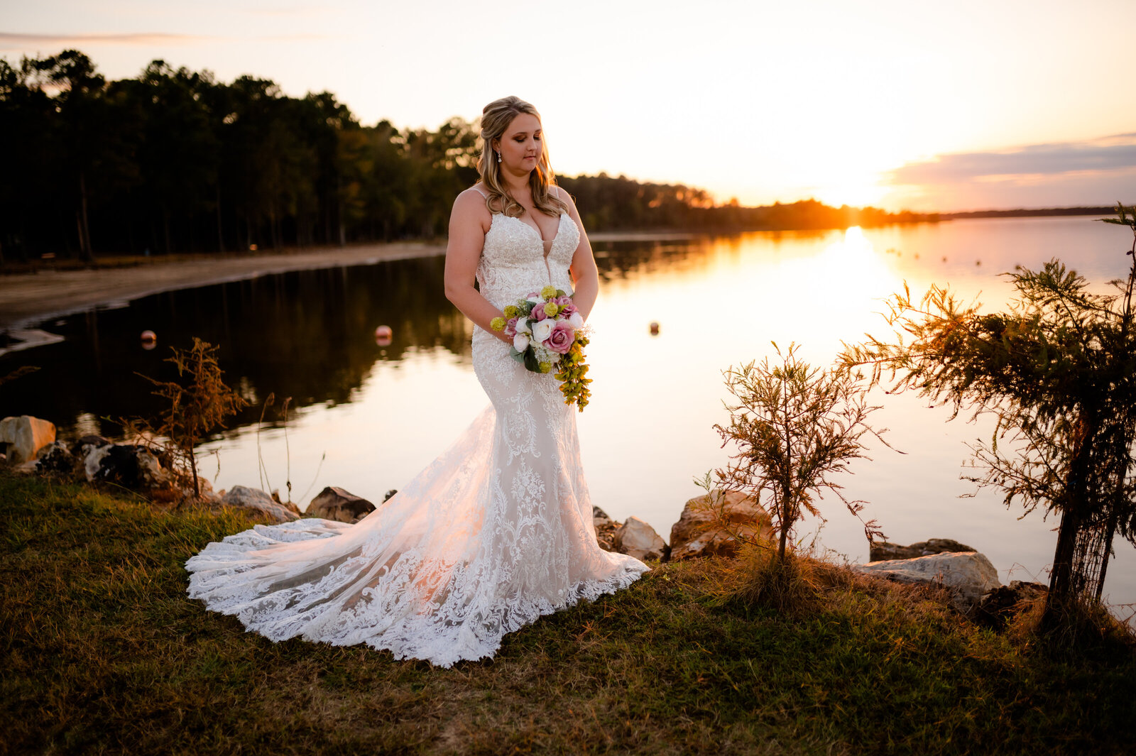 bride standing on a lake as the sun sets behind her and reflects to the bride and her lace dress