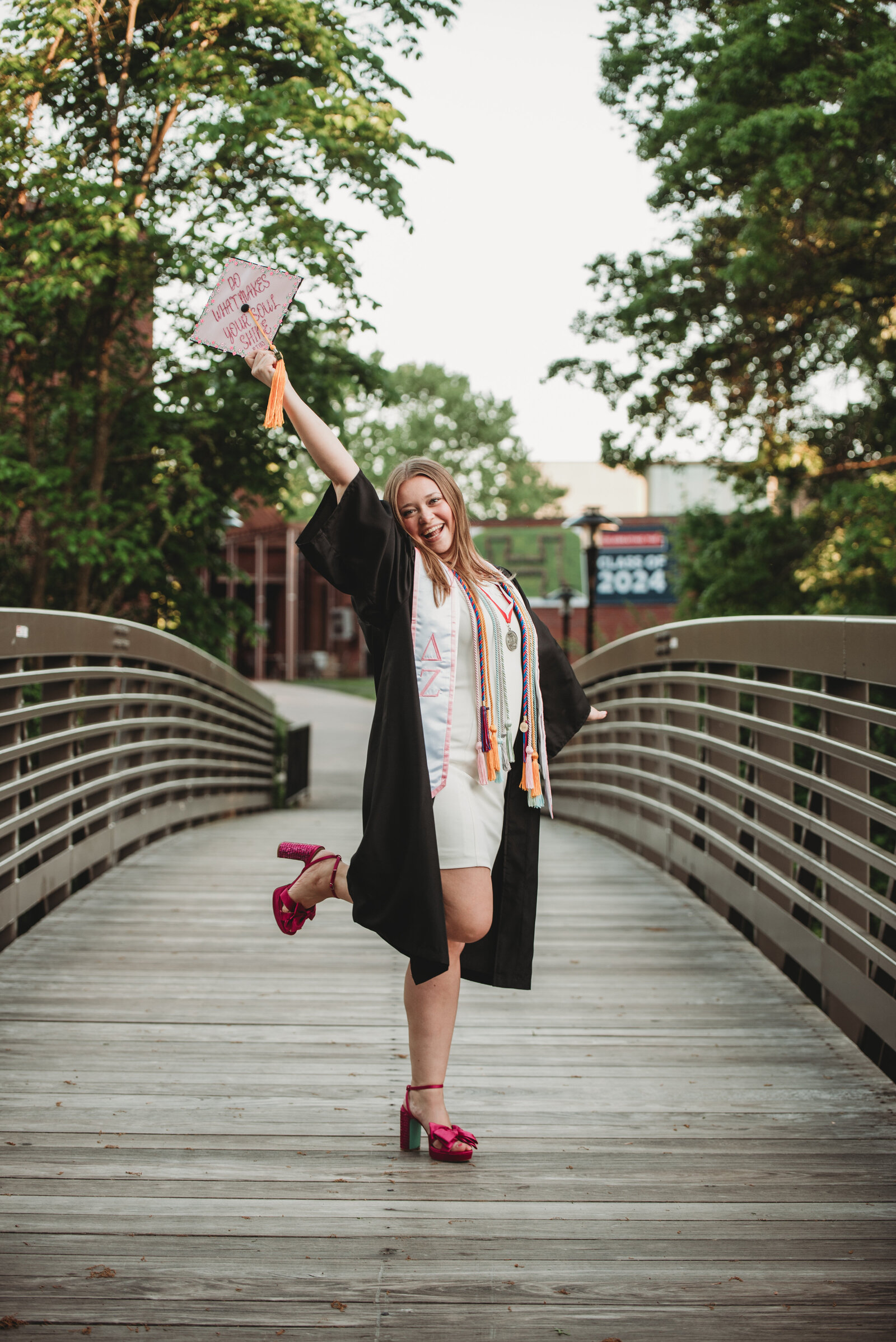 A University of Hartford graduate in a black cap and gown poses joyfully on a campus bridge, holding her decorated graduation cap high in celebration. Captured during a Connecticut college graduation photography session by Nicole Mele Photography, showcasing achievement and excitement.