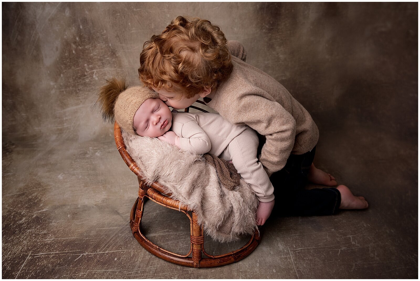 Young toddler kissing new baby brother on the  side of the face, who is sleeping in a papasan chair.