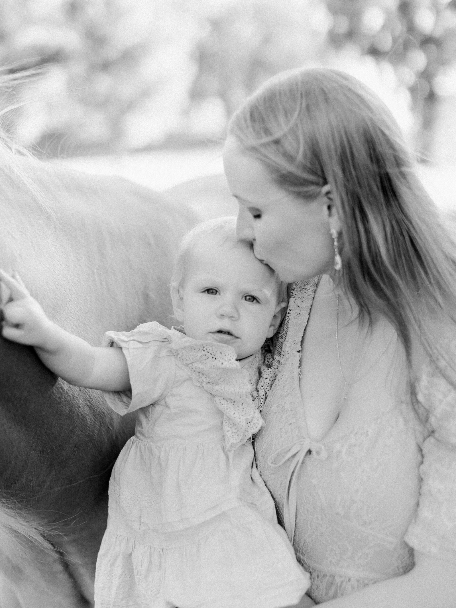 Closeup emotional black and white motherhood portrait of a mom holding her on year old on her hip and kissing her head while baby pets  their palomino horse on a horse farm by NH newborn photographer Fieldstone Studio.