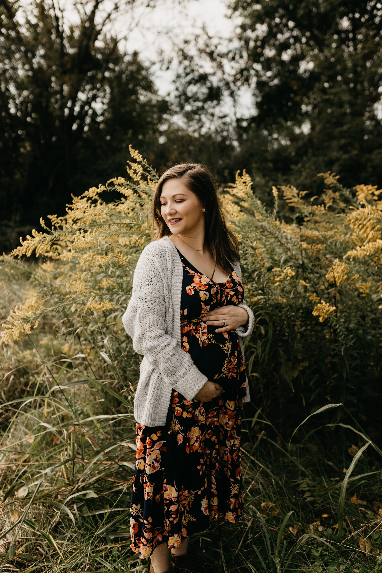 A pregnant woman in a floral dress stands in front of a bush of yellow flowers
