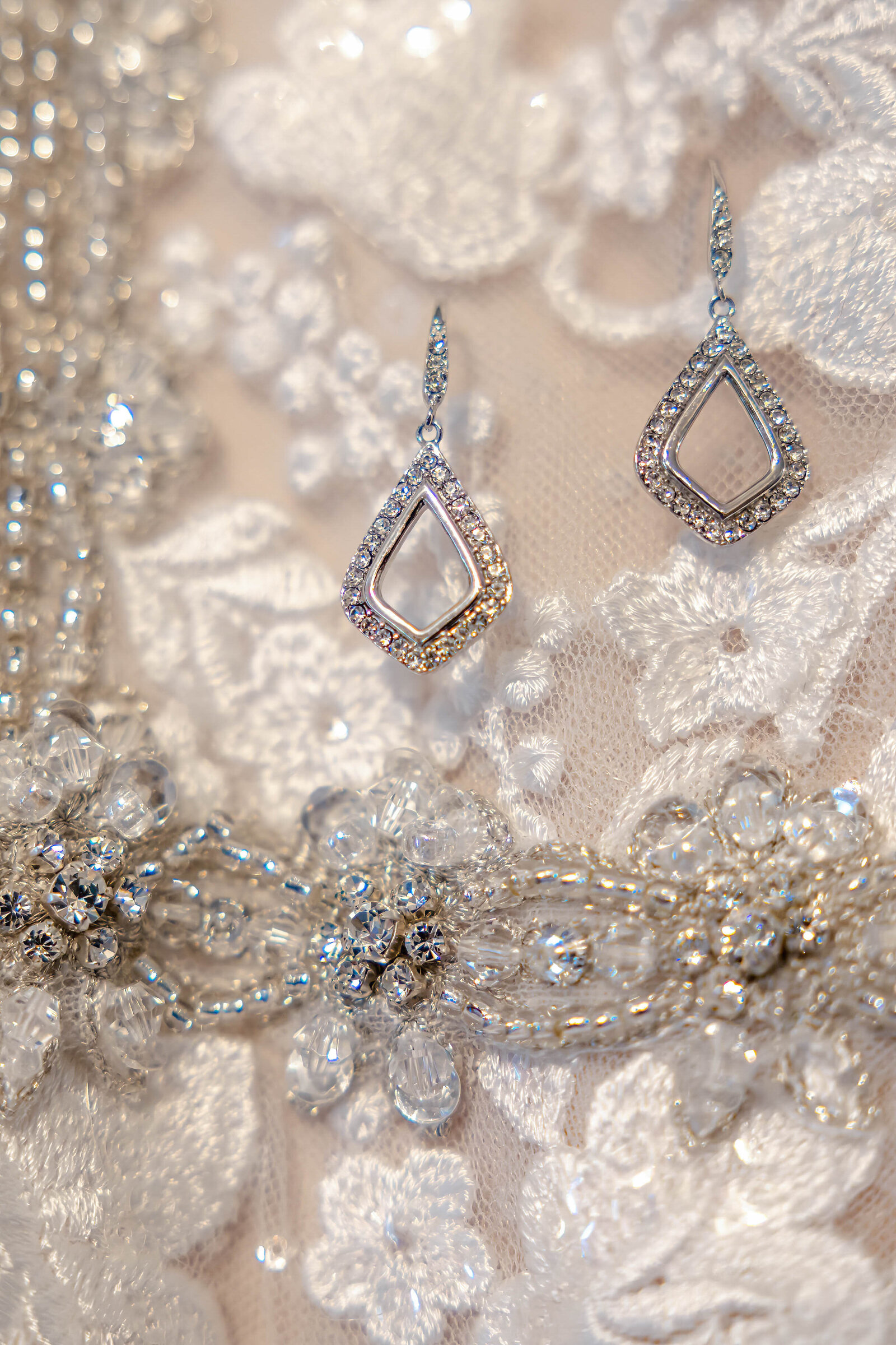 Close-up of sparkling diamond teardrop earrings and a vintage-inspired belt displayed on a lace wedding dress, showcasing intricate bridal details for a Oak Island, NC wedding.