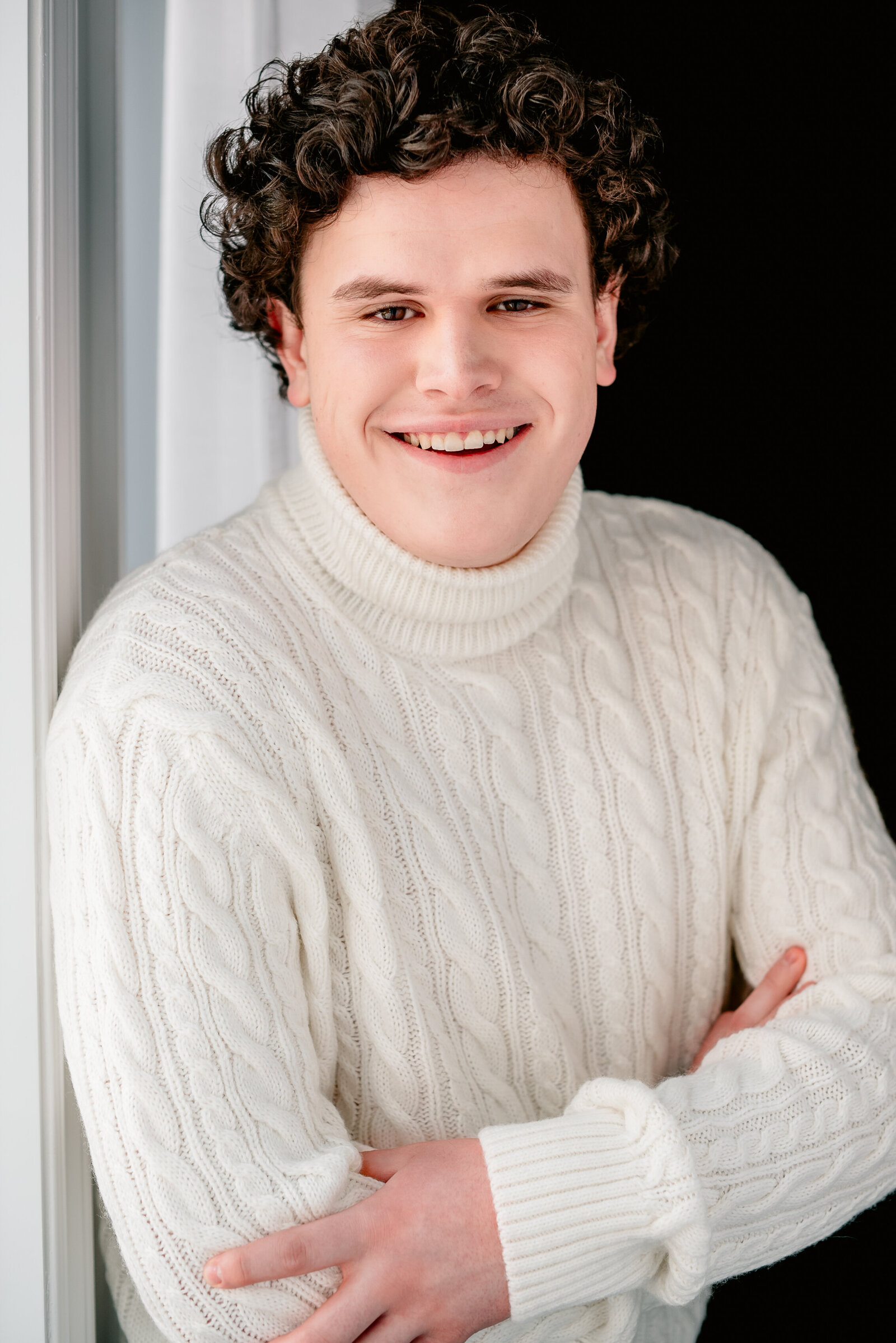 Headshot of a young man poses in front of a window with arms crossed wearing an ivory cable-knit sweater