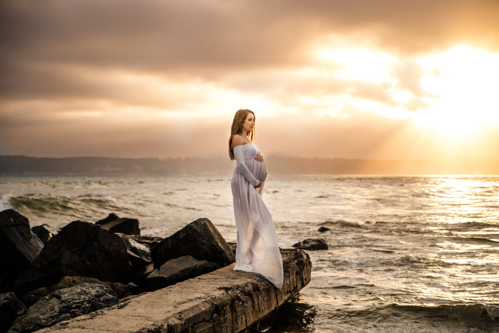 Maternity Photographer, a pregnant woman stands on ocean rocks in a maternity gown