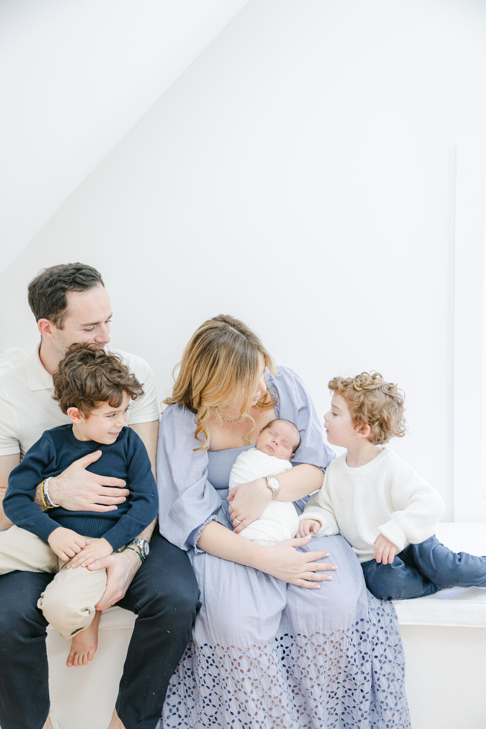 A candid family moment during a lifestyle newborn session in Fairfield County, CT, featuring parents and their three young children sitting together in a bright, airy room. Nicole Neira Photography captures the connection, joy, and tender sibling interactions in a soft, organic setting.