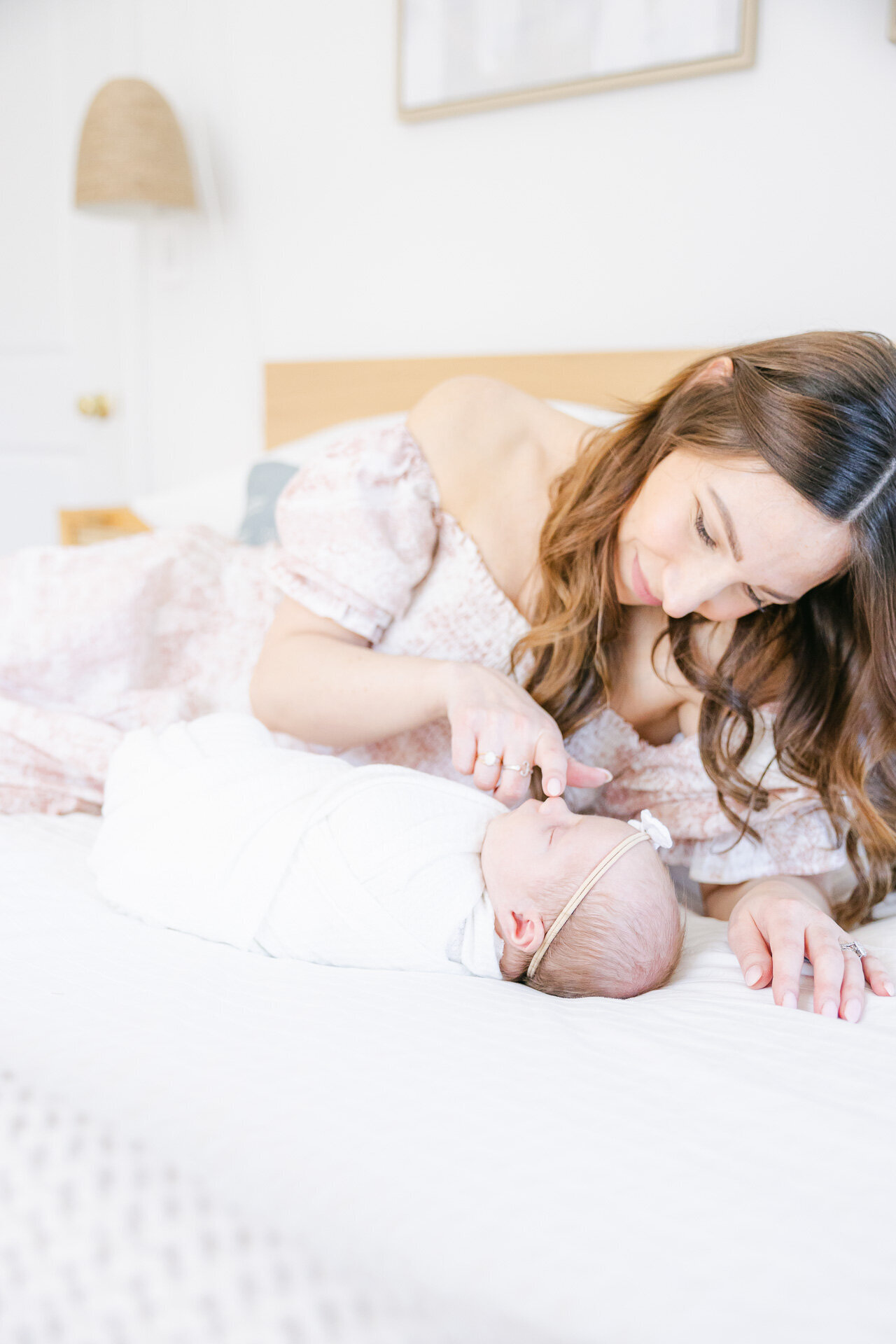 A mother in a floral off-the-shoulder dress gently lays beside her swaddled newborn on a cozy white bed, smiling tenderly as she touches the baby’s nose. Captured in natural light and soft tones, this image embodies the warmth and wonder of early motherhood—perfect for families who want to remember the tiniest moments in the sweetest way.