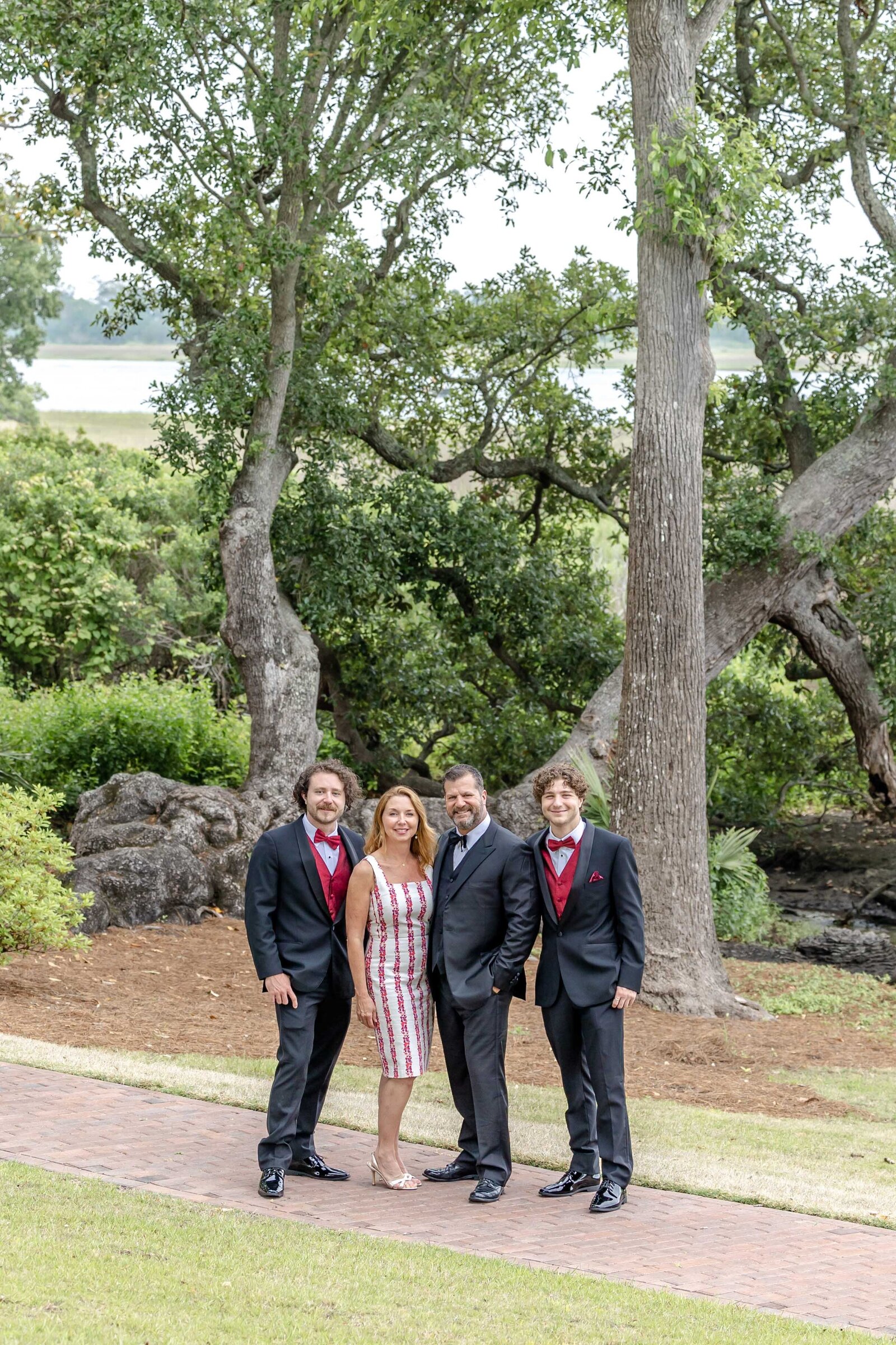 A family poses elegantly on a garden path, with men in suits and a woman in a patterned dress. The lush greenery and trees provide a sophisticated backdrop, ideal for professional family photography in NC, highlighting elegance and style.