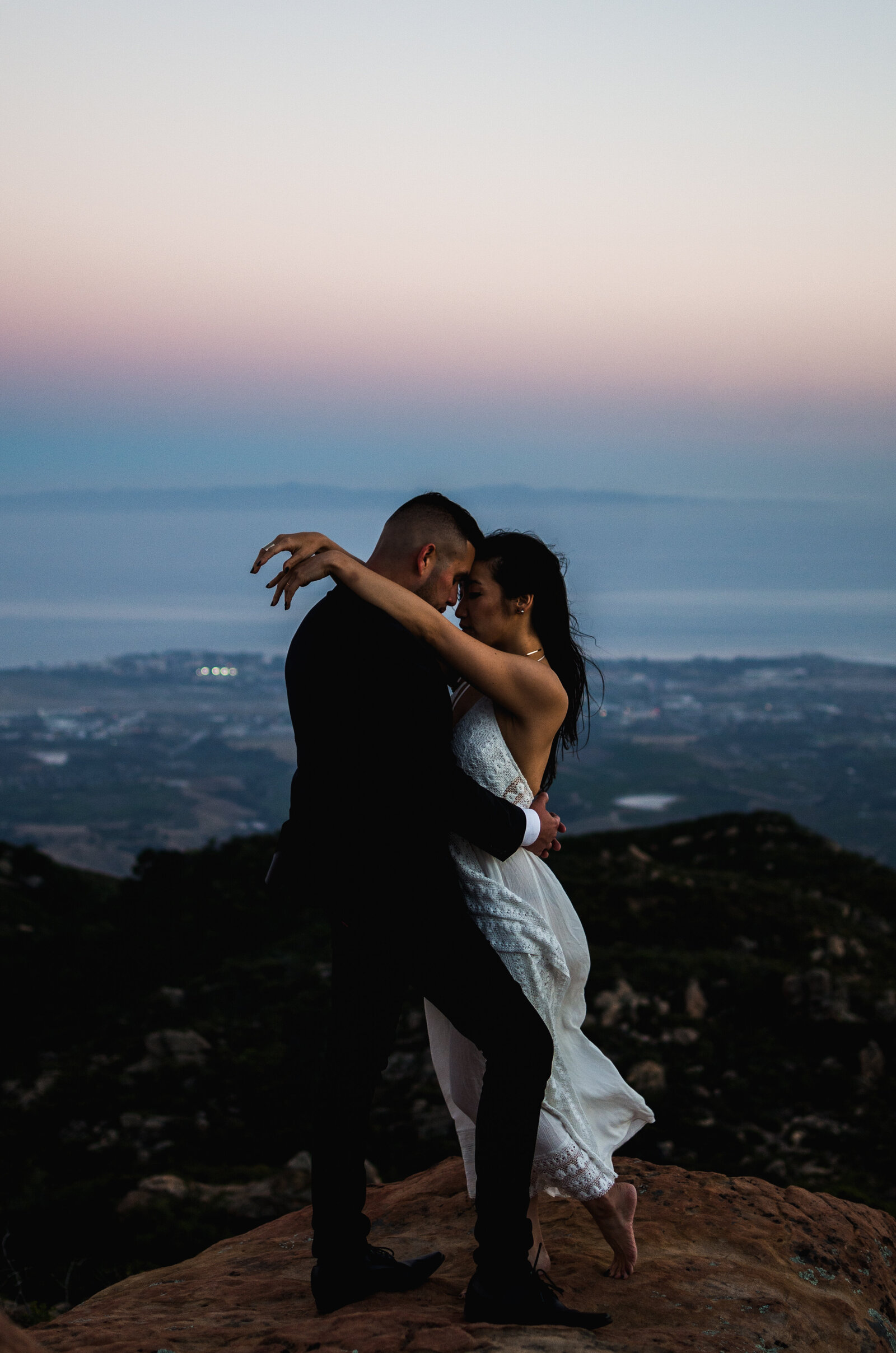 Couple holding each other on a rock on top of the mountain, with the city in the background.
