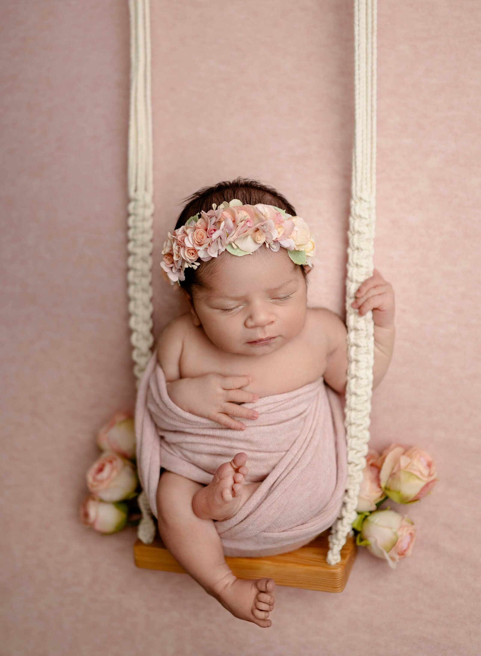 Baby girl swaddled in pink wrap and holding onto swing with roses.