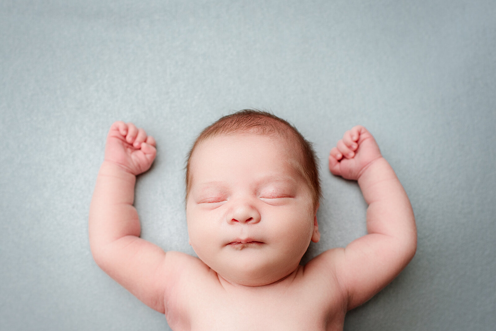 Light blue backdrop with newborn boy laying on back hands above head in St. Johns, FL.