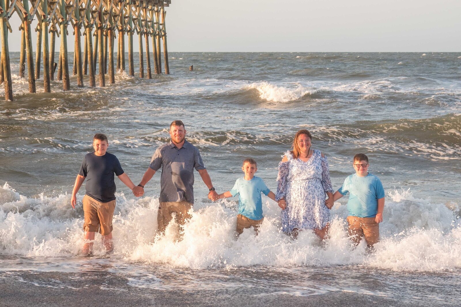 A family of five stands in the ocean waves near a pier, dressed in blue and khaki outfits. The lively surf and wooden structure create a captivating scene, ideal for destination family photographer NC, capturing adventurous and playful moments.