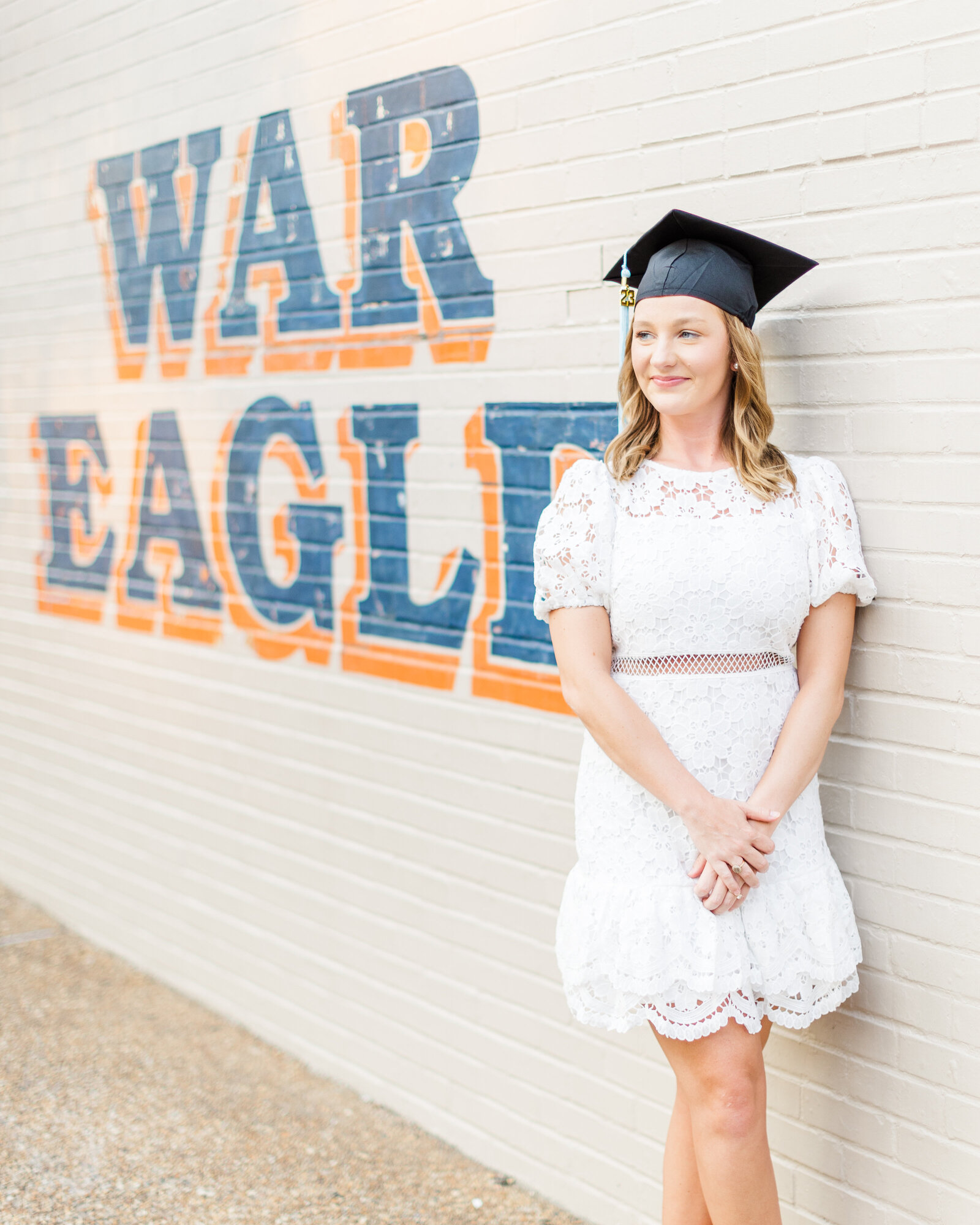 Shelby posing against a brick wall with 'War Eagle' painted in bold letters, wearing her graduation attire, captured by Abby Bell Photography.