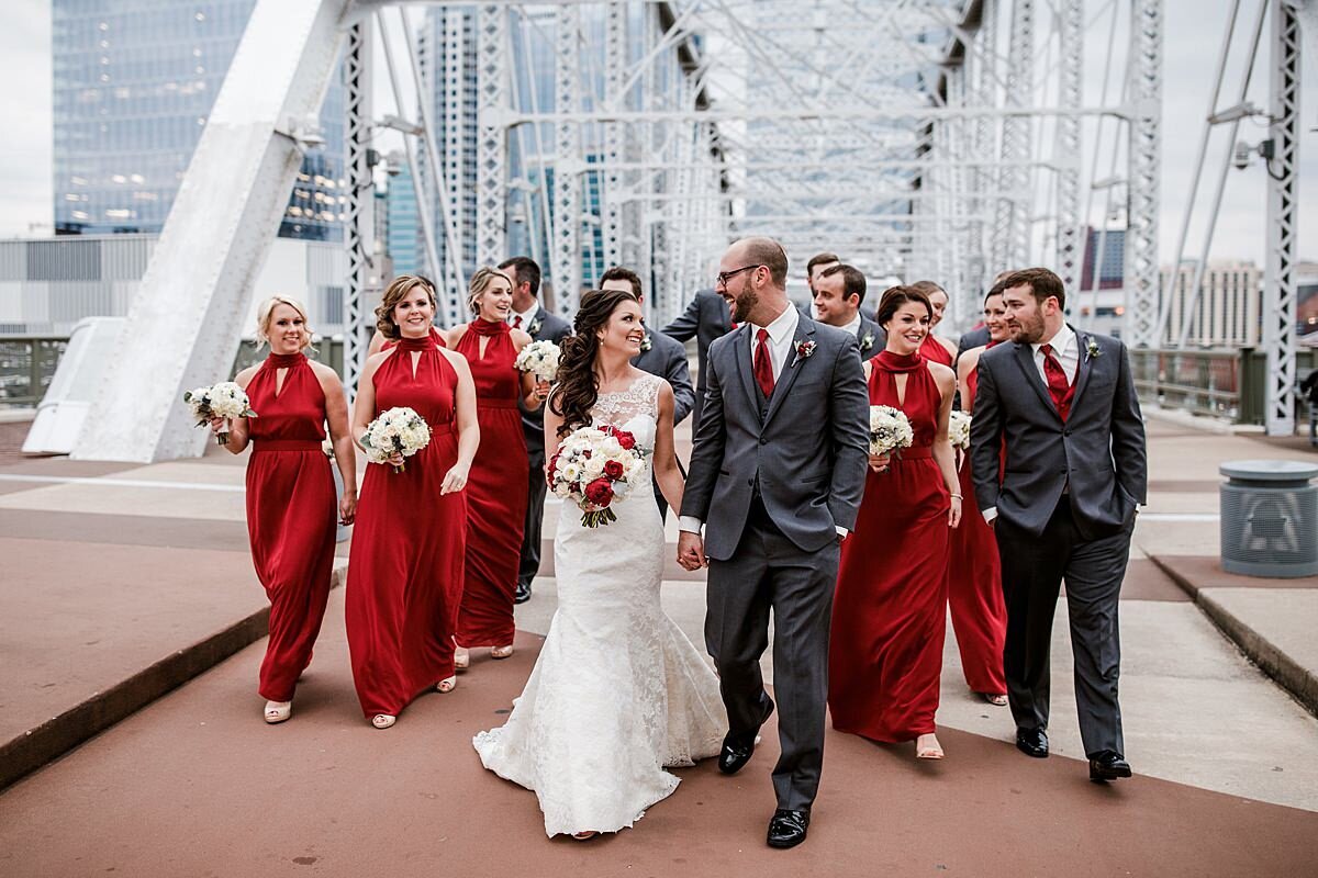 The bride in a lace trumpet gown and the groom in a charcoal gray suit lead the bridal party dressed in navy and red along the pedestrian bridge in Nashville.