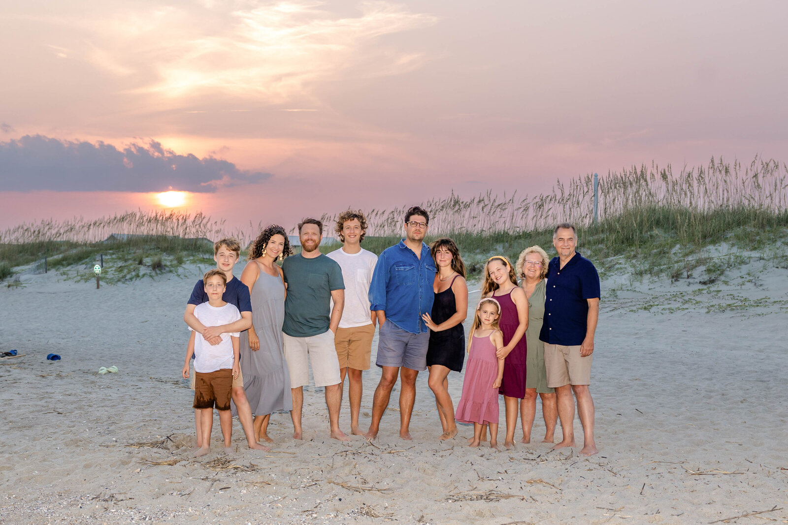 A family poses on sandy dunes at sunset, dressed in casual summer attire. The setting sun and pastel sky create a warm and serene atmosphere, ideal for professional family photography in NC, capturing intimate and relaxed moments.